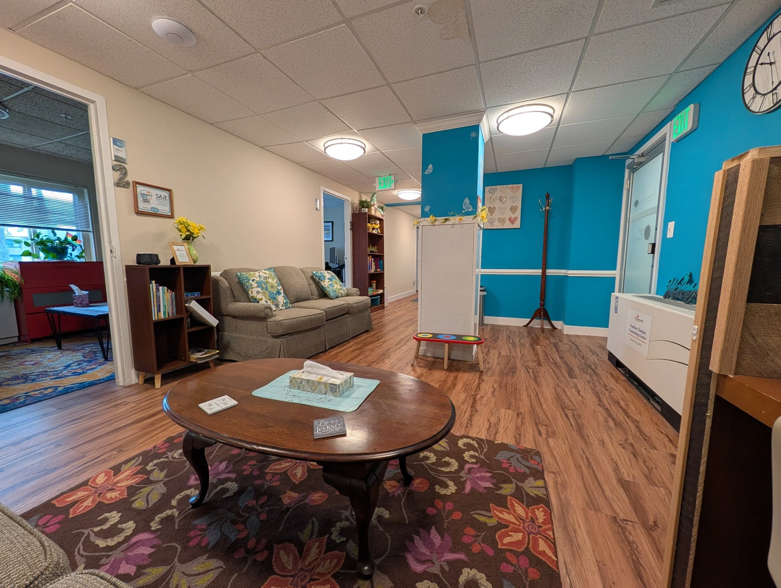 A waiting area with a wooden coffee table on a colorful floral rug, a beige sofa with blue and yellow pillows, bookshelves, and wall decorations including a clock and butterflies.