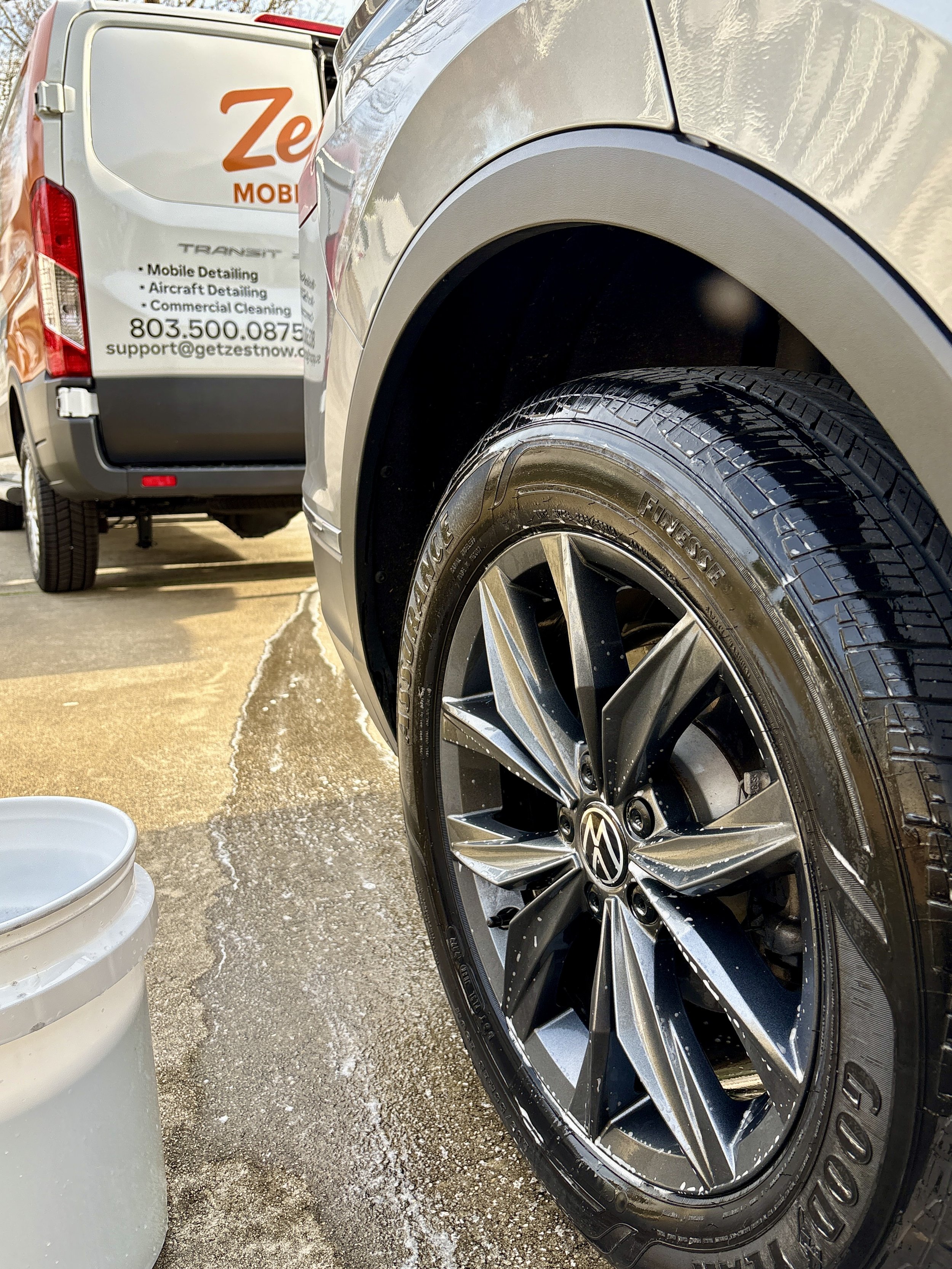 Close-up of a car's front wheel with a chrome rim and a Goodyear tire. A white and grey van parked nearby has a sign advertising a mobile detailing business, including contact information and services such as aircraft detailing and commercial cleaning.