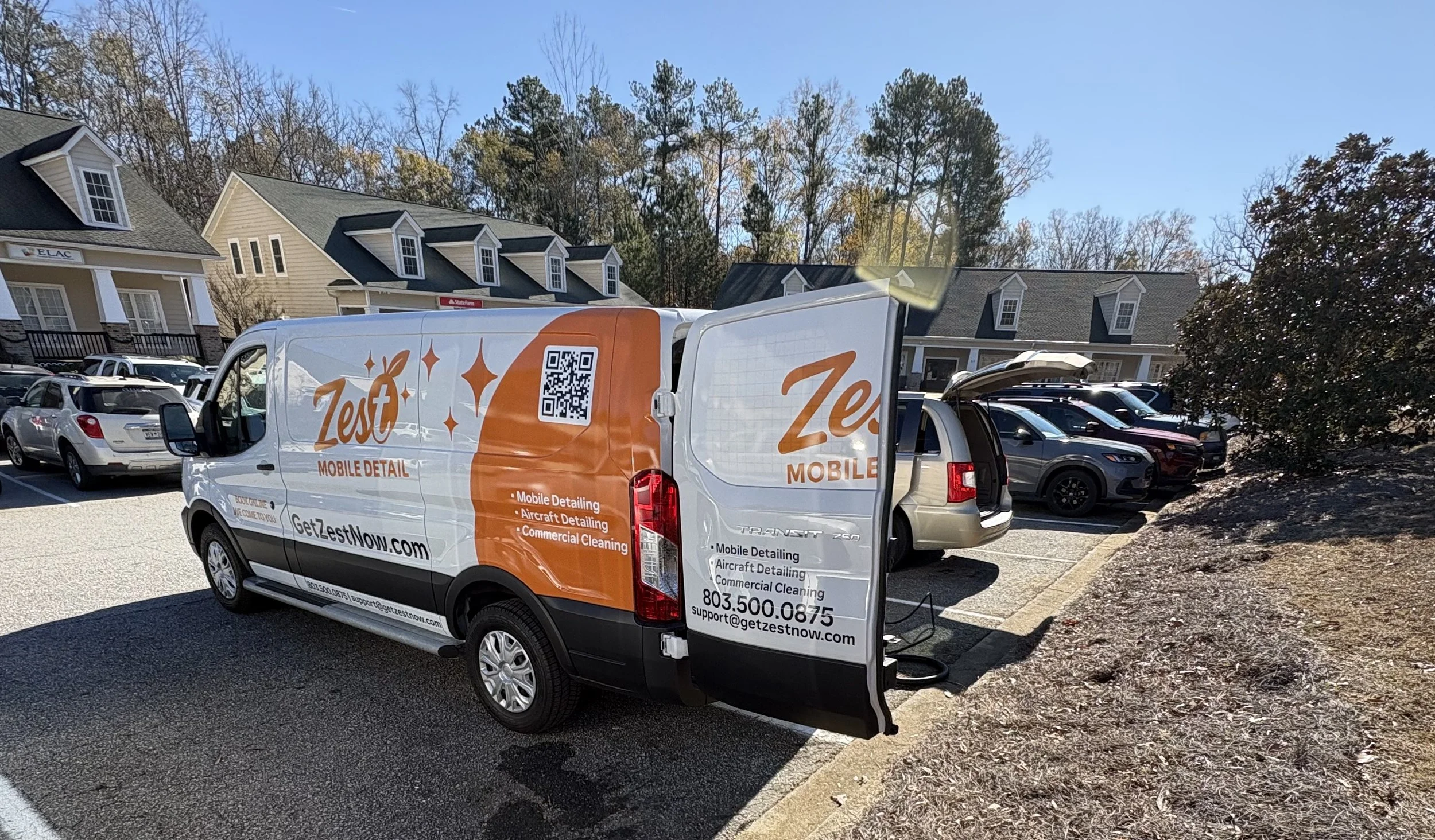 A Zest Mobile Detail service van parked in a parking lot with its rear doors open. Several other vehicles are parked nearby and residential buildings can be seen in the background. The van has branding and contact information for Zest Mobile Detailin