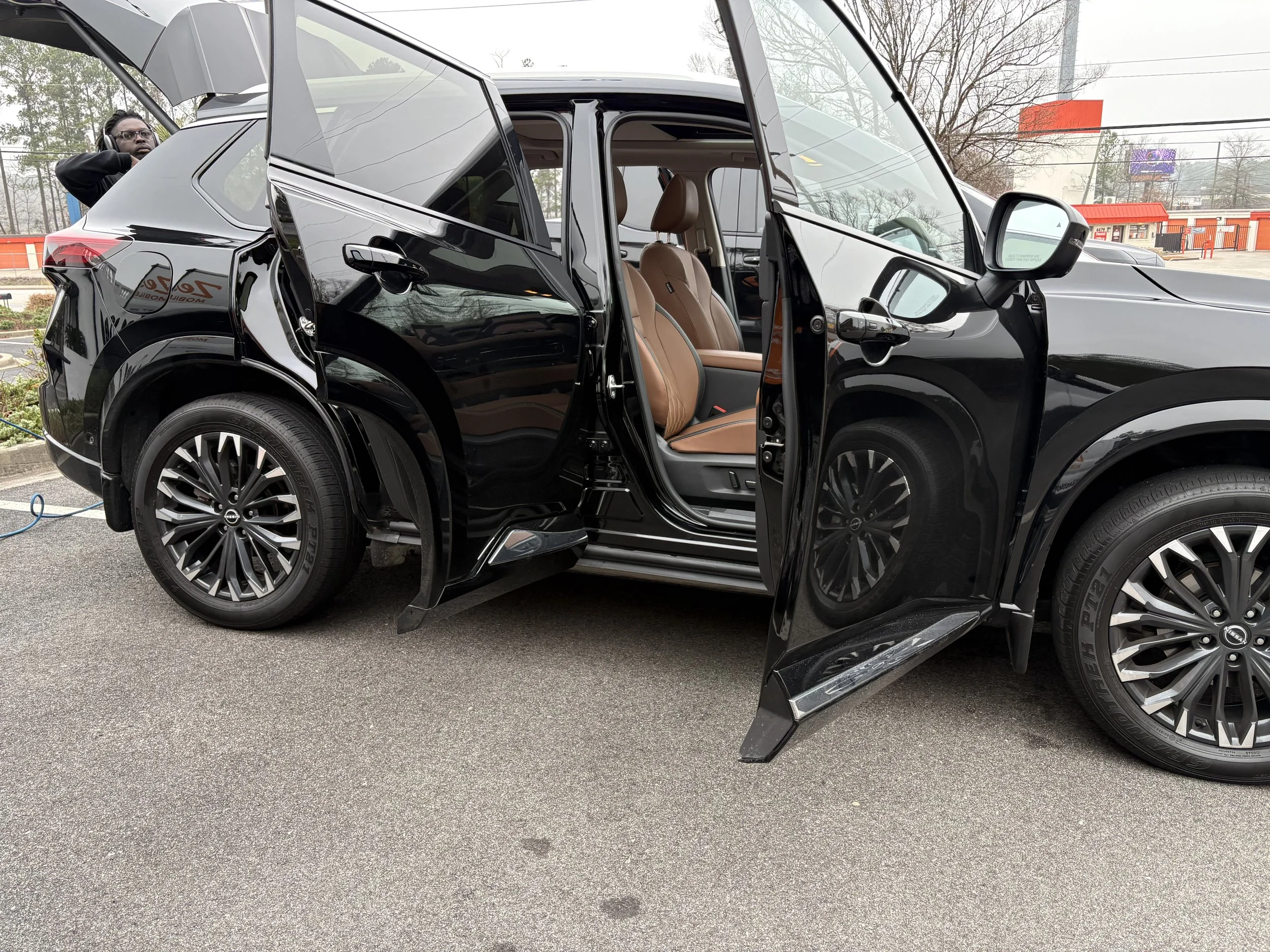 A black SUV with its doors open, showing tan interior seats, parked on a paved lot with a person standing nearby and a background of trees and commercial buildings.