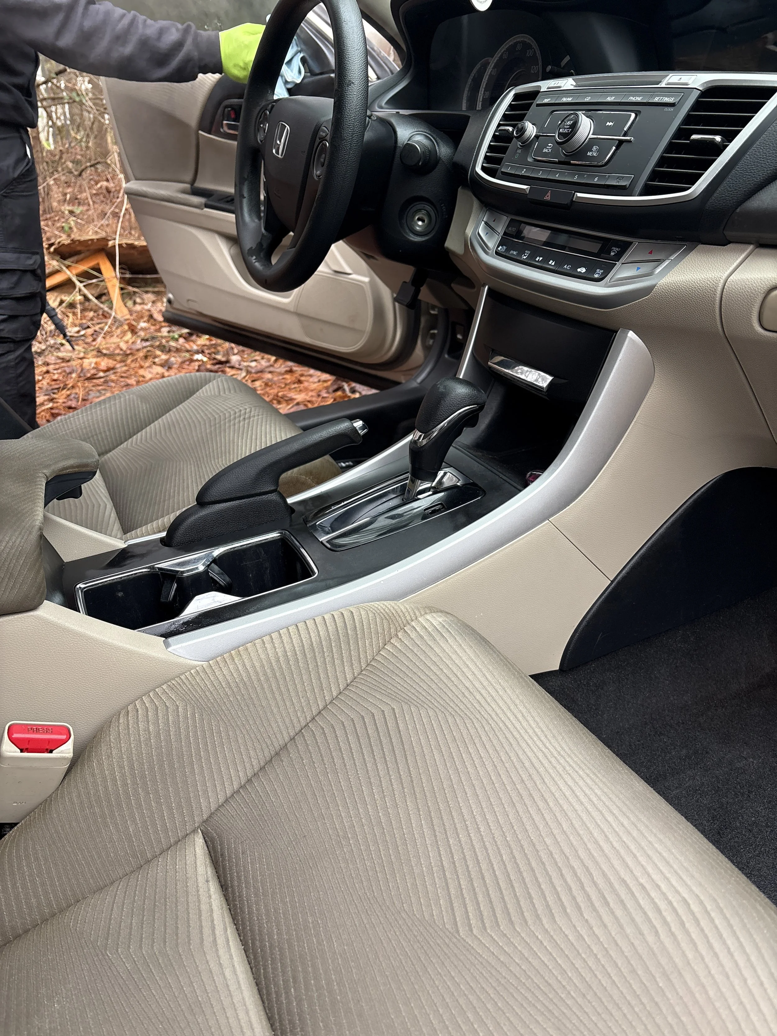 The interior of a car showing front seats, gear shift, steering wheel, dashboard controls, and an open driver-side door with a person standing outside near a wooded area.