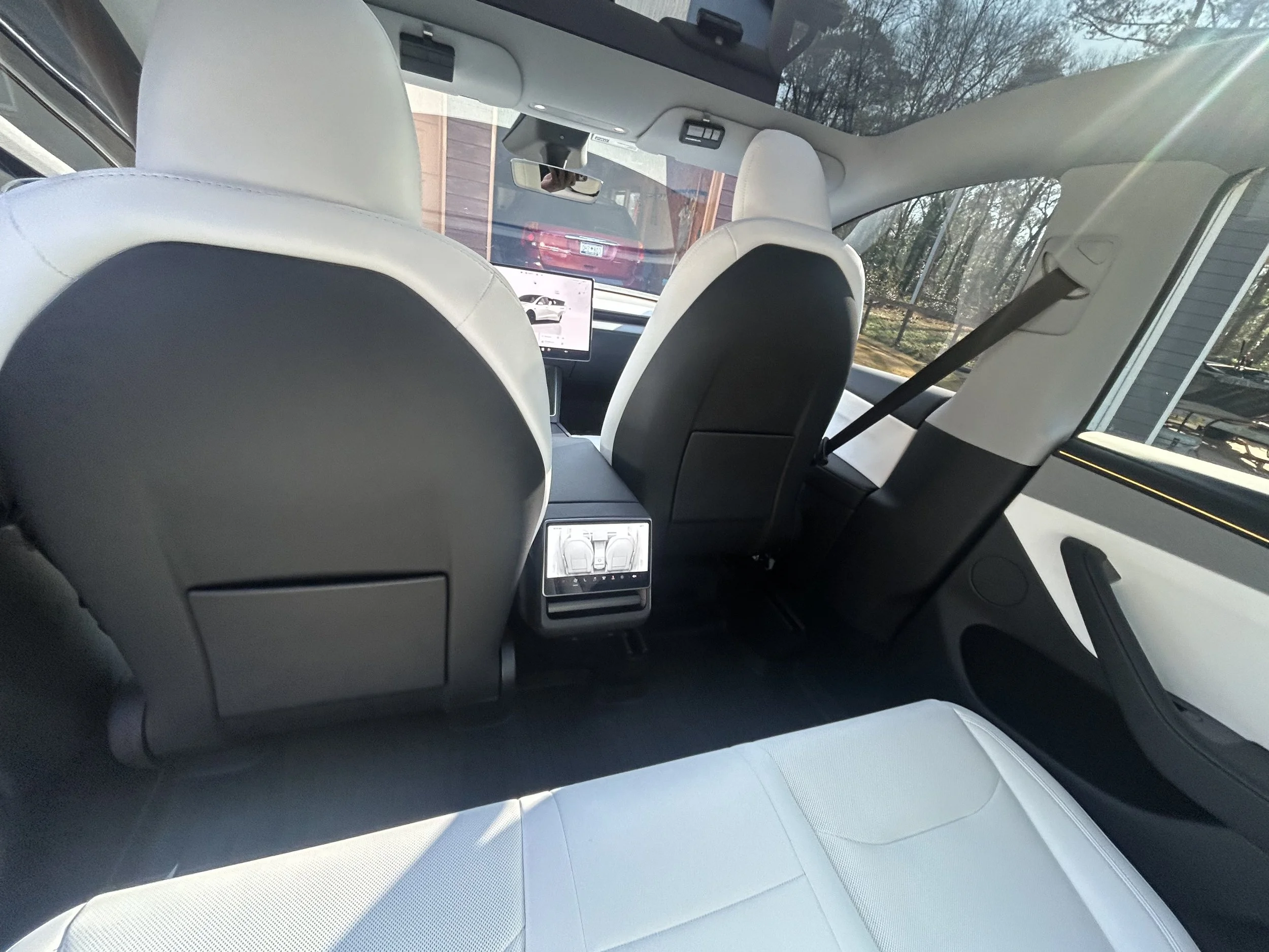 Inside an electric vehicle, front and rear seats visible with seatbacks, a central rear-seat screen, rear air vents, and a digital climate control panel. The view through the windshield shows parked cars and trees outside.