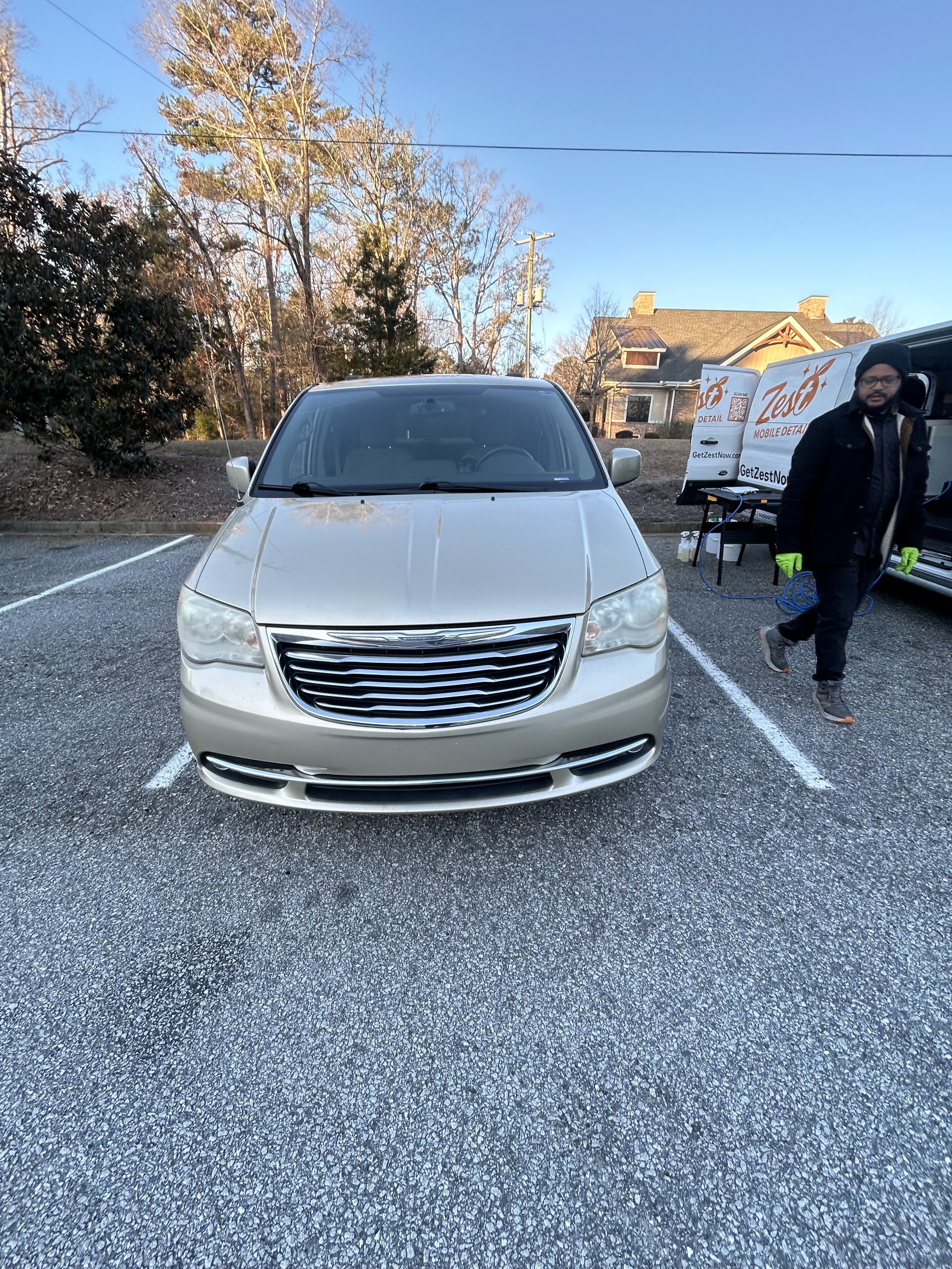 Front view of a beige Chrysler minivan parked in a parking lot with a man in black coat and yellow gloves walking past.