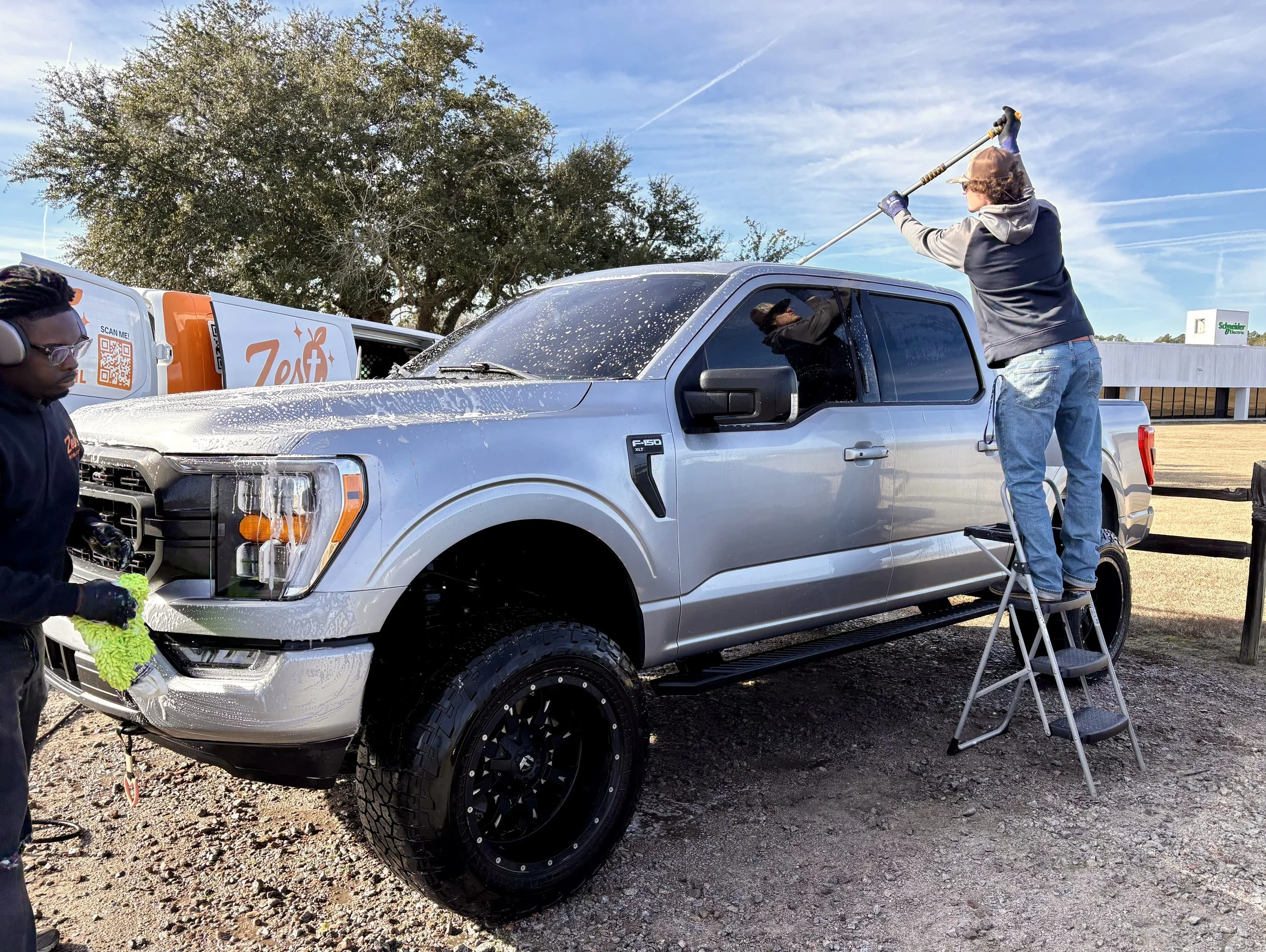 Two people washing a silver pickup truck outdoors; one person cleaning the truck's windshield from a stepladder, another with a hose or sponge near the front of the vehicle; a van with a logo and QR code in the background.