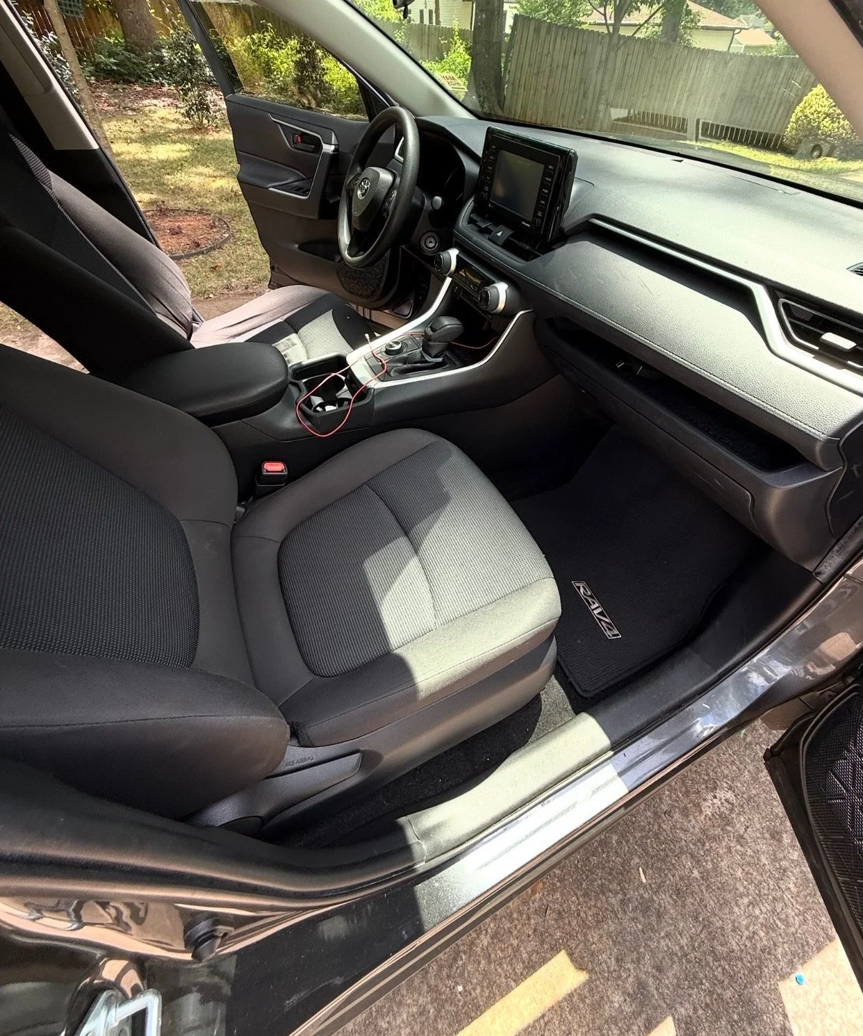 Interior of a car showing the driver's seat, dashboard, steering wheel, center console with gear shift, and a touchscreen display. The car is parked outdoors, with some greenery visible through the windows.