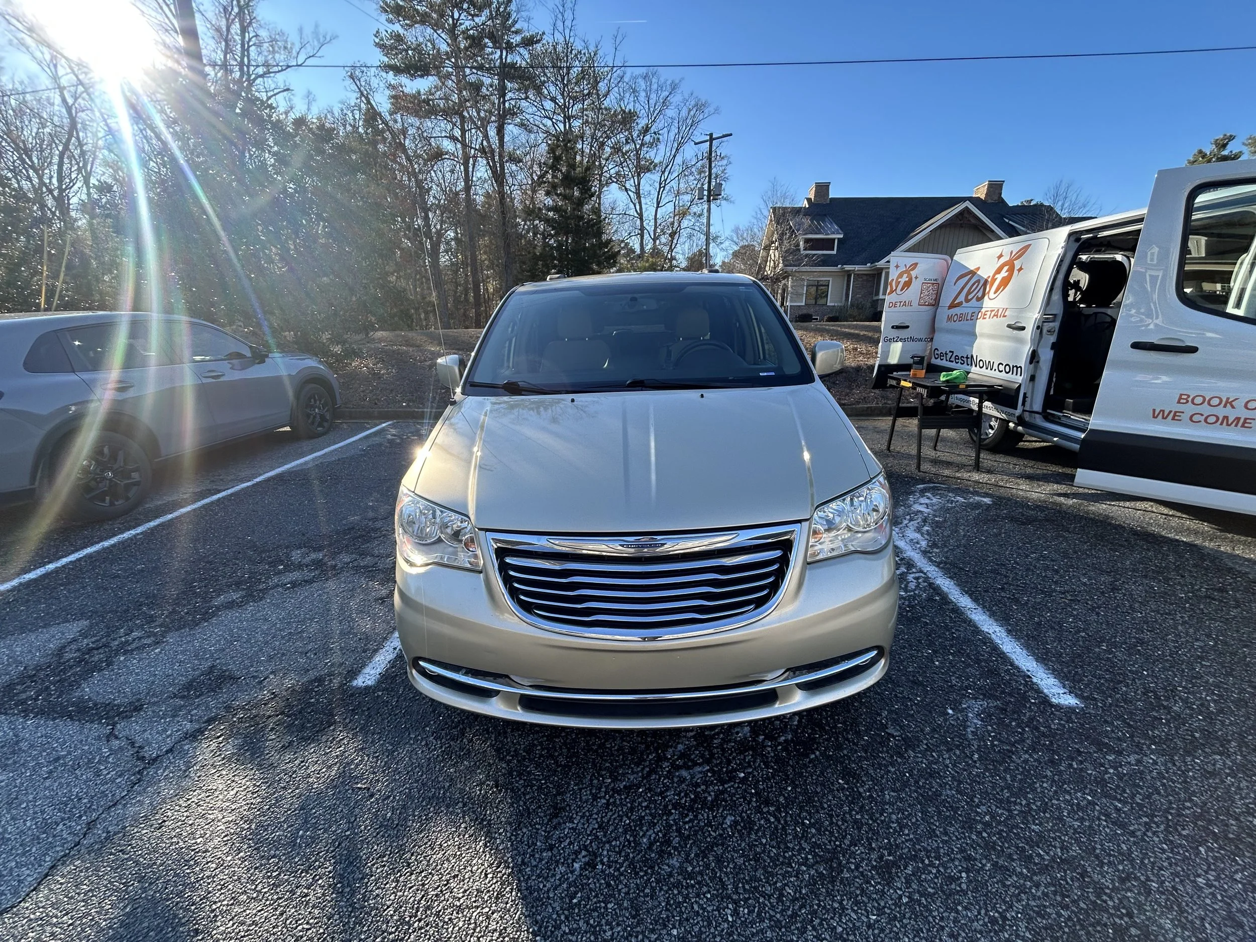 Silver Chrysler minivan parked in a parking lot on a sunny day, with a white and gray house and a mobile detailing truck in the background.