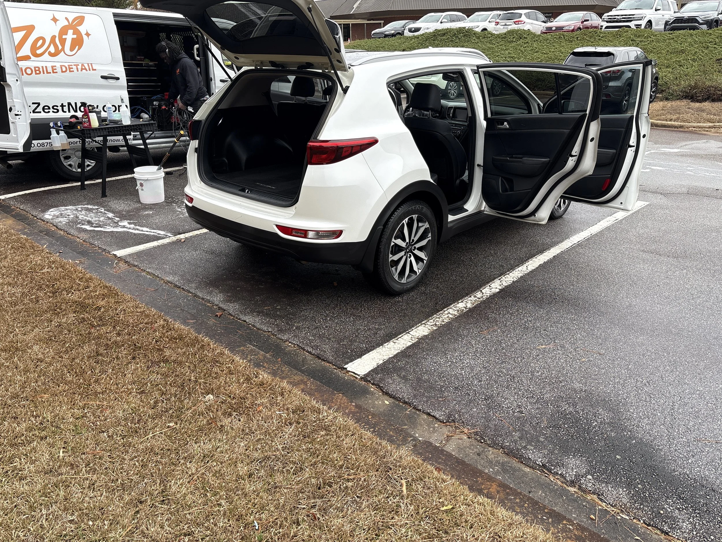 A white car parked in a parking lot with its rear hatch open and front passenger doors open. A mobile detailing van is parked next to it, with a worker cleaning or detailing the vehicle.