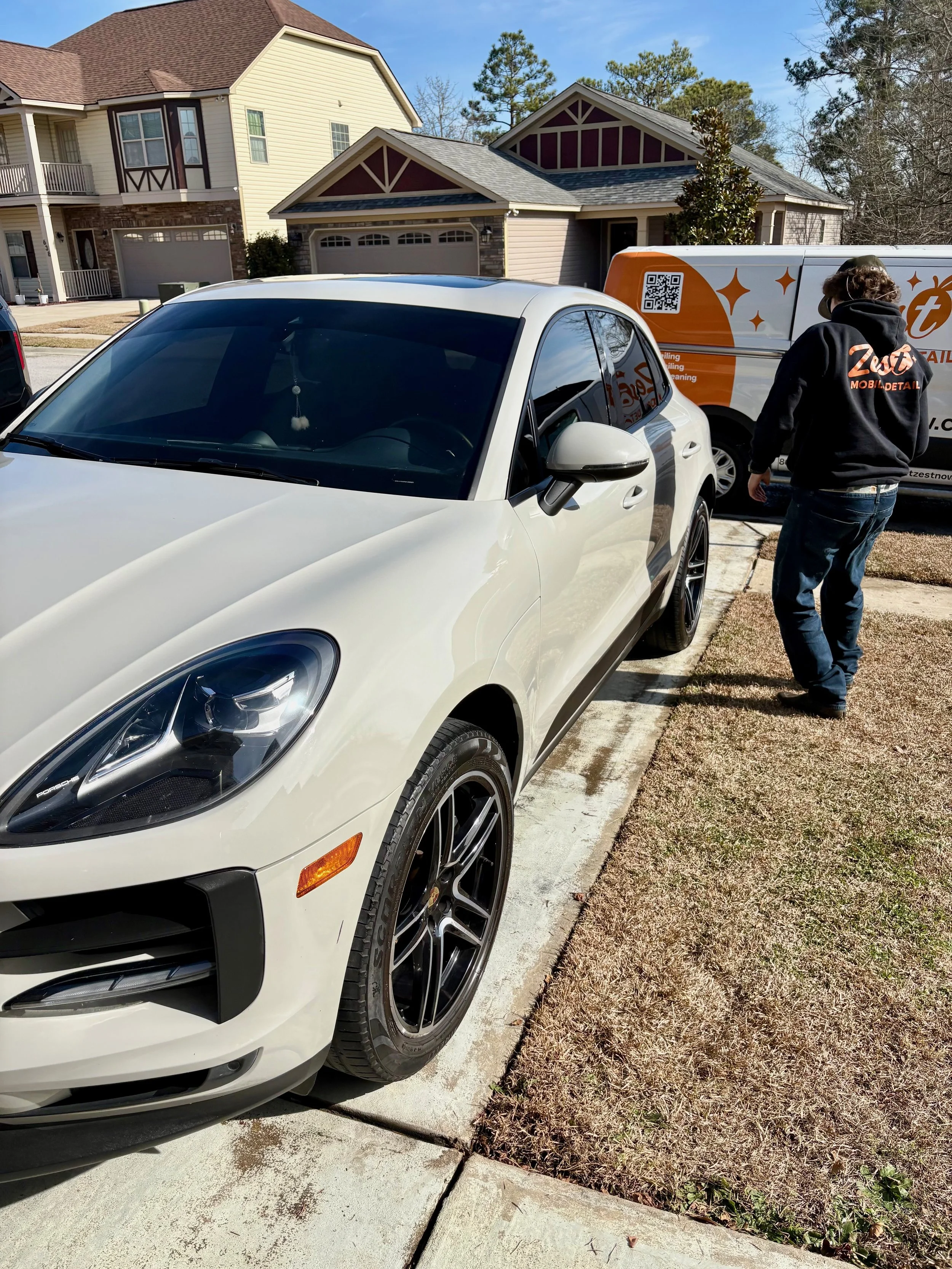 White Porsche Macan parked on a driveway next to a person wearing a black hoodie with orange text. In the background are houses and a delivery van with orange and white branding.