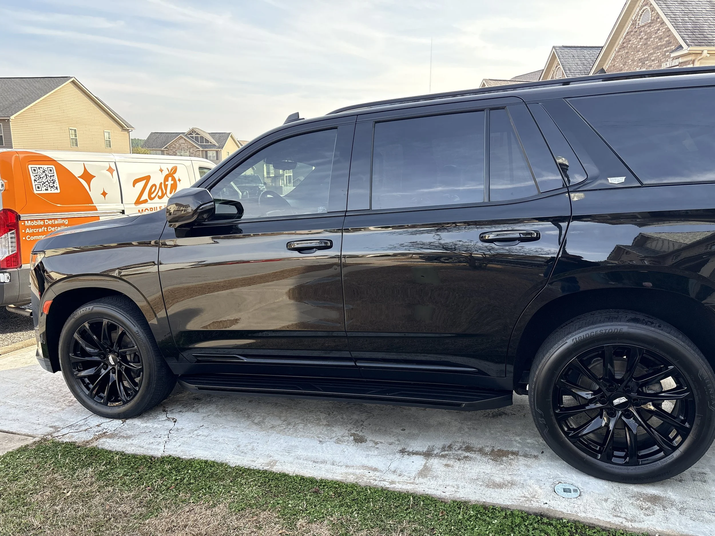 A black SUV parked on a concrete driveway with houses in the background and an orange and white vehicle in the background.