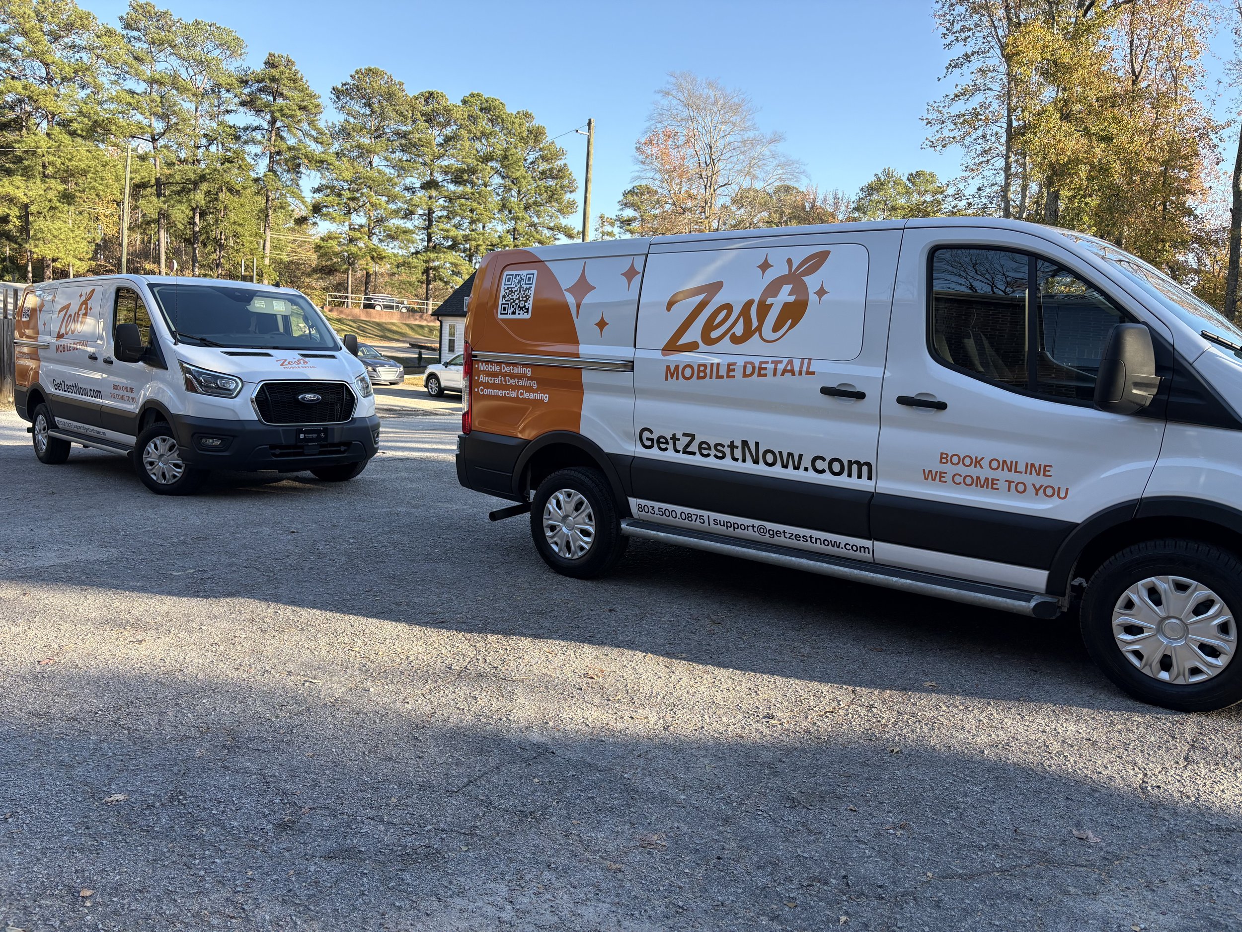 Two Zest Mobile Detailing service vans parked outdoors on a gravel surface. The vans have the Zest logo and contact information, with a background of trees and a blue sky.