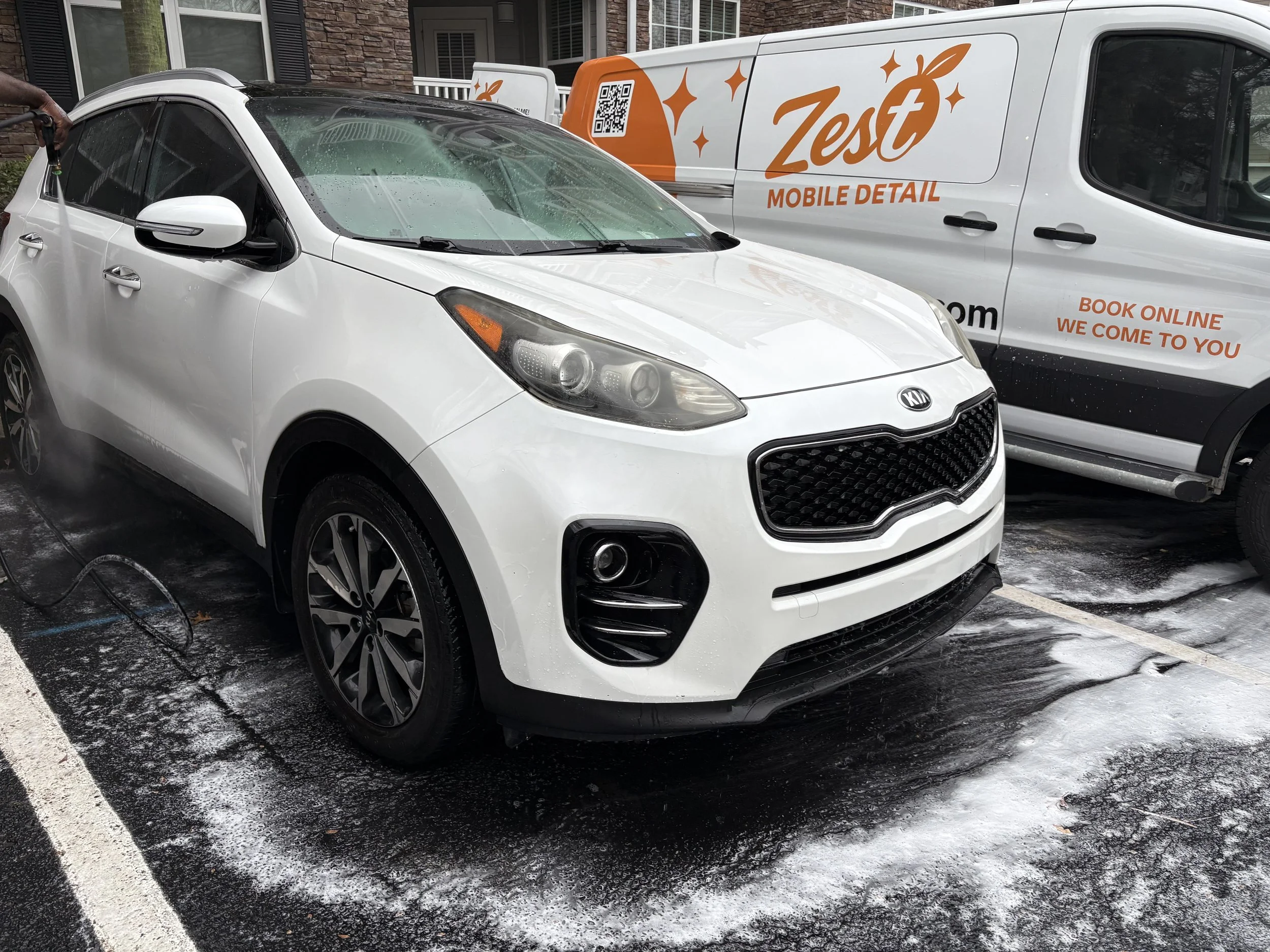 A white Kia SUV being washed with soap and water at an outdoor car wash station. Foam is visible on the ground and the vehicle.