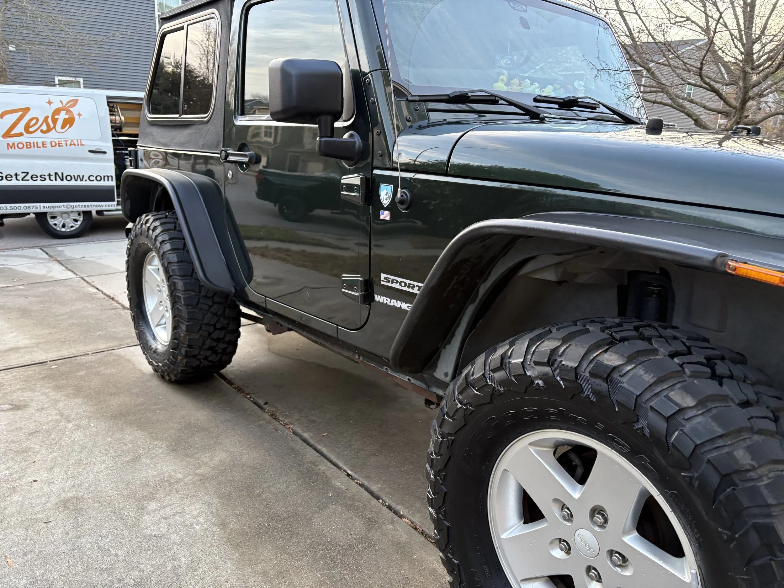 A black Jeep Wrangler Sport parked on a driveway with large off-road tires, next to a white van with orange and black branding for Zest Mobile Detail.