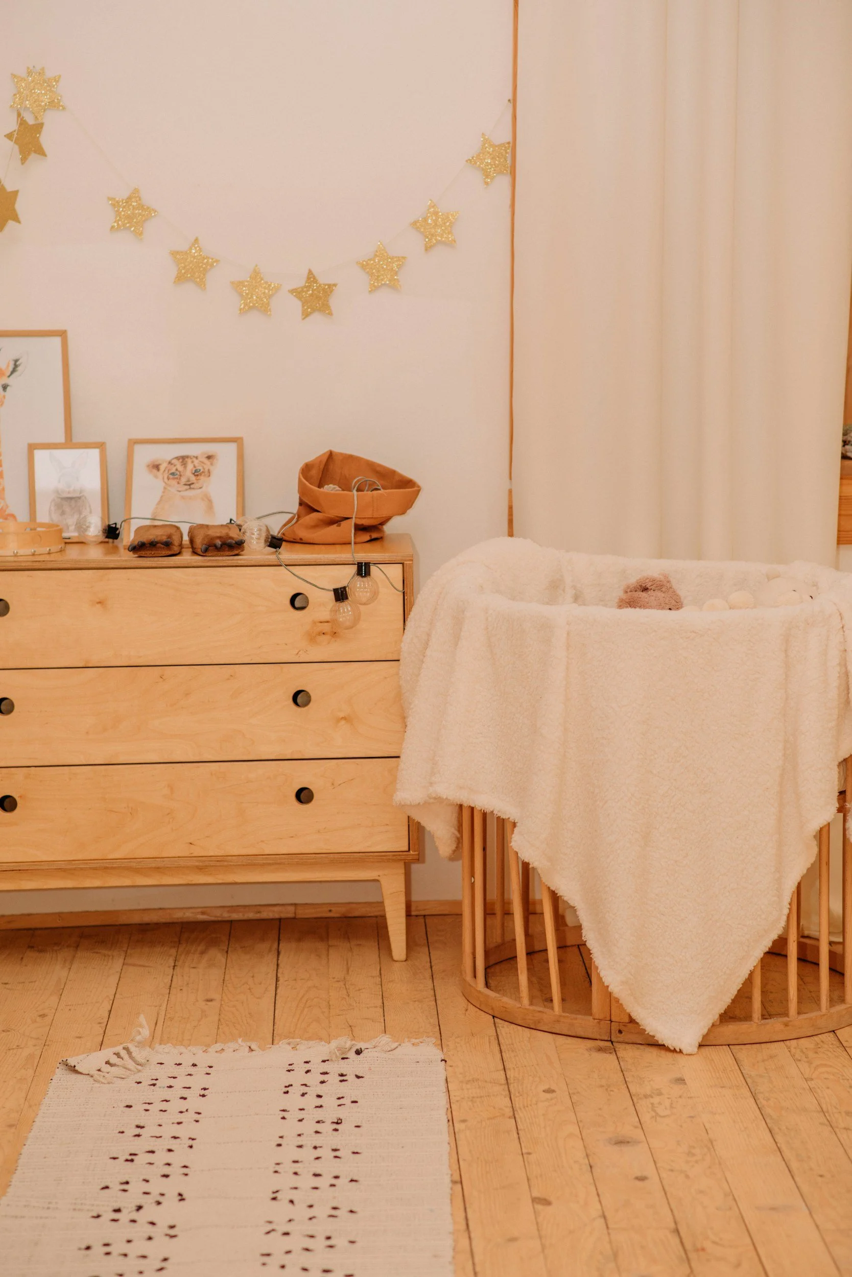 A cozy nursery corner with a light wood dresser, decorated with framed animal-art prints, a string of gold star garland, and a string of light bulbs. Next to the dresser is a round wooden baby bassinet draped with a soft cream blanket, and a small striped rug on the wooden floor.
