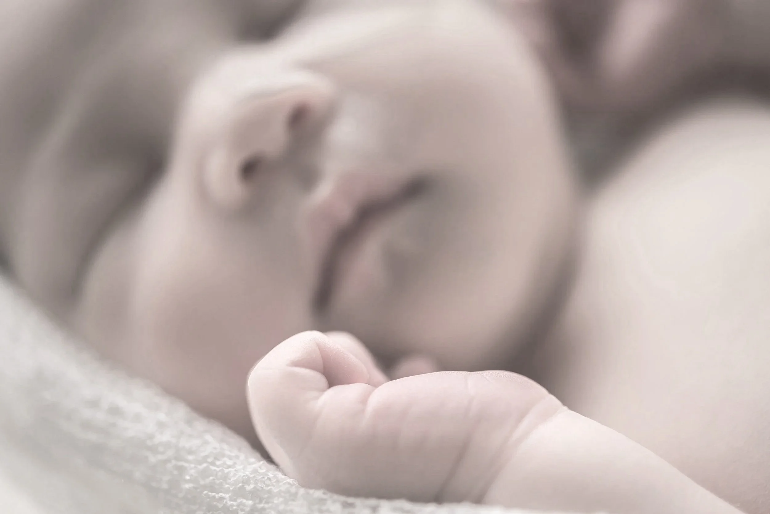 Close-up of a newborn baby lying down with a blurred face in the background, showing a tiny hand curled into a fist and soft skin.