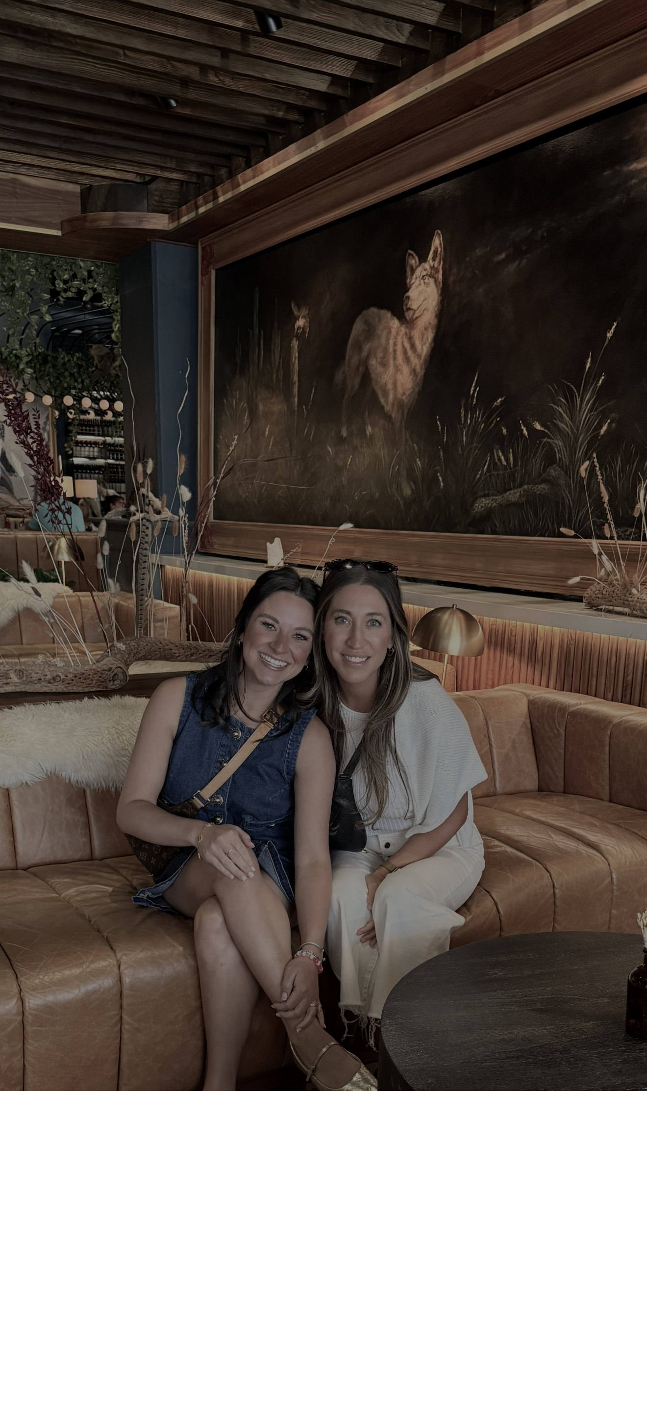 Two women sitting on a tan leather couch inside a warmly lit restaurant with an art piece of a mountain lion on the wall behind them.