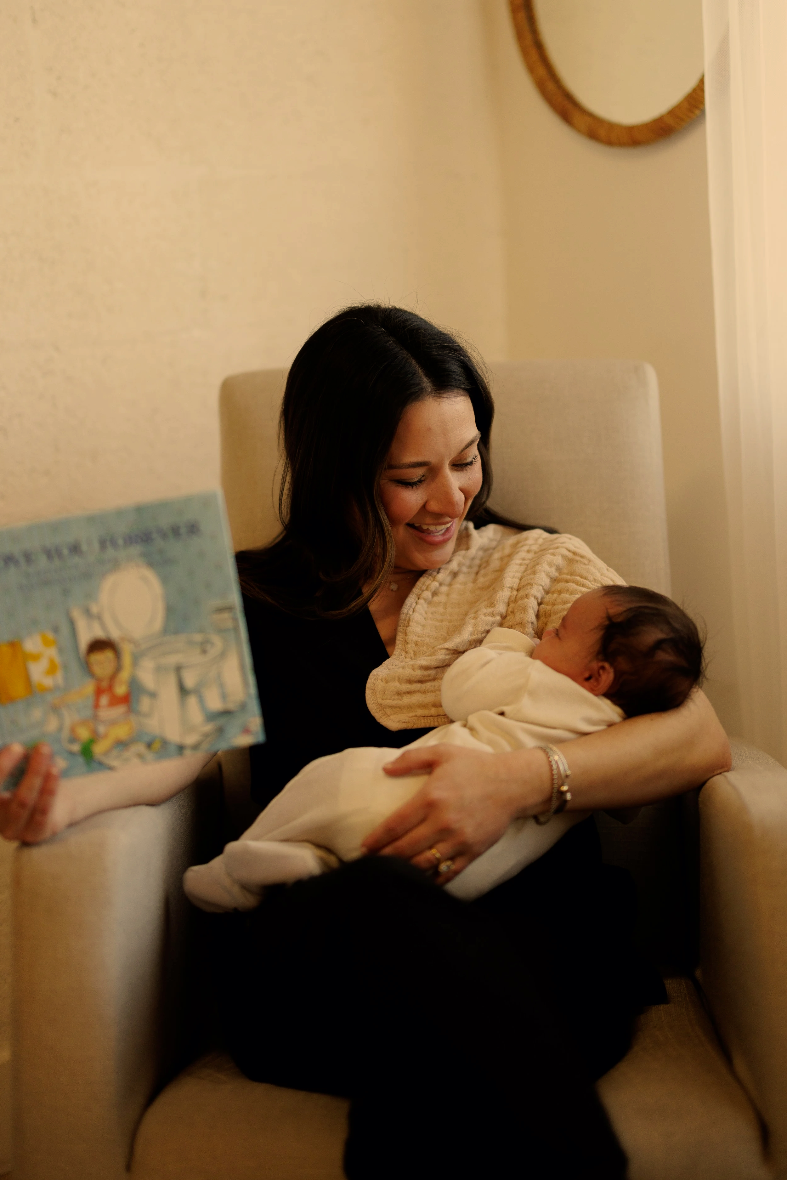 A woman holding a baby and smiling at him while sitting in a beige armchair, with a child holding a illustrated children's book in the foreground.