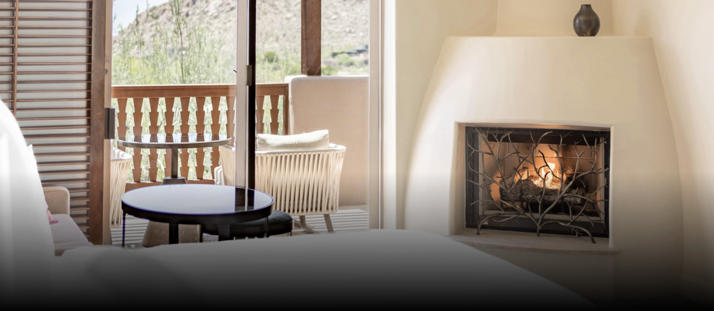 Interior of a cozy room with a white fireplace showing a lit fire, a small black side table, a lamp with a black shade, and an open balcony with card chairs and a view of greenery outside.