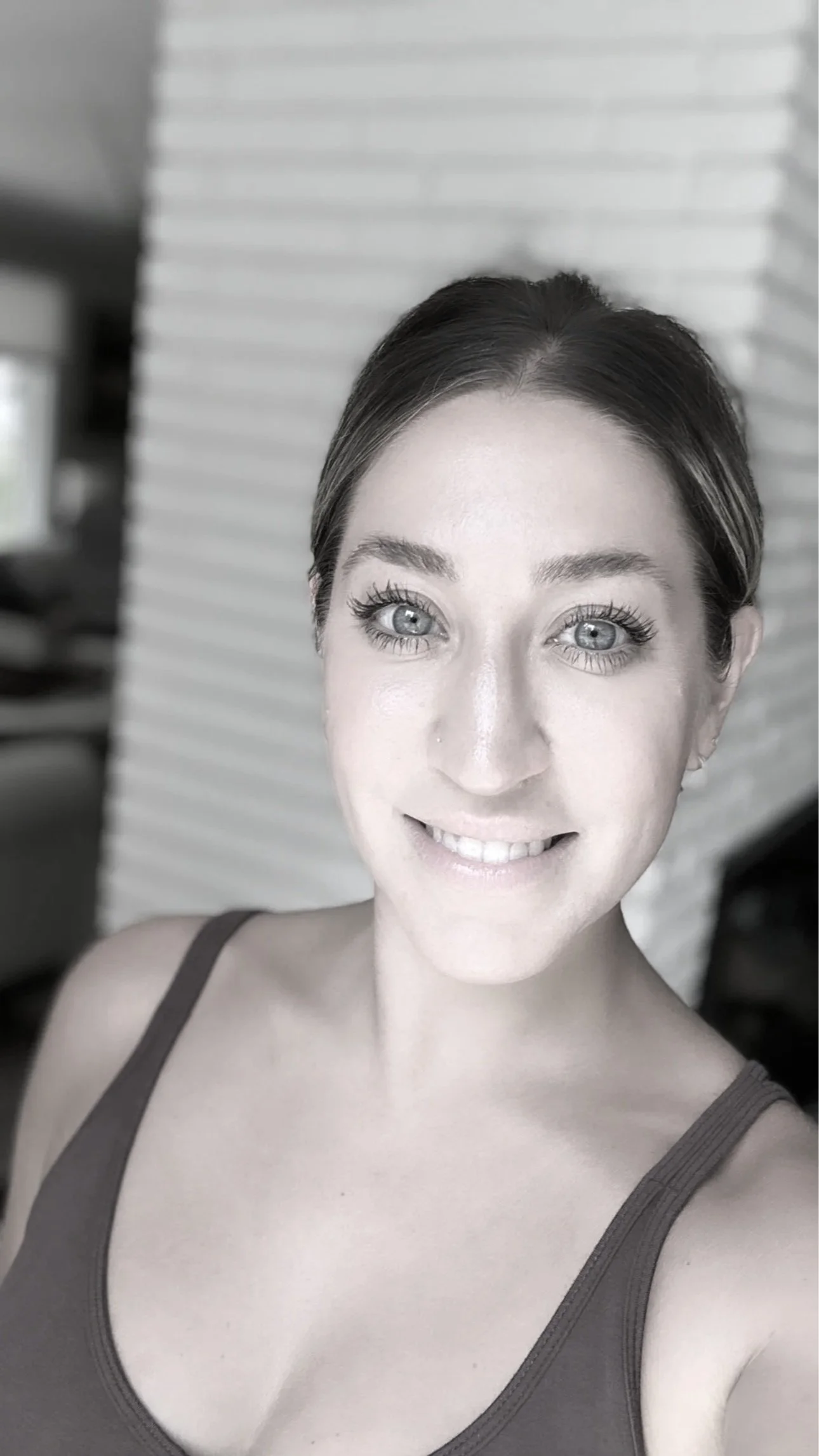 A woman with blue eyes and dark hair in a bun, smiling at the camera, wearing a tank top, standing indoors in front of a brick wall.