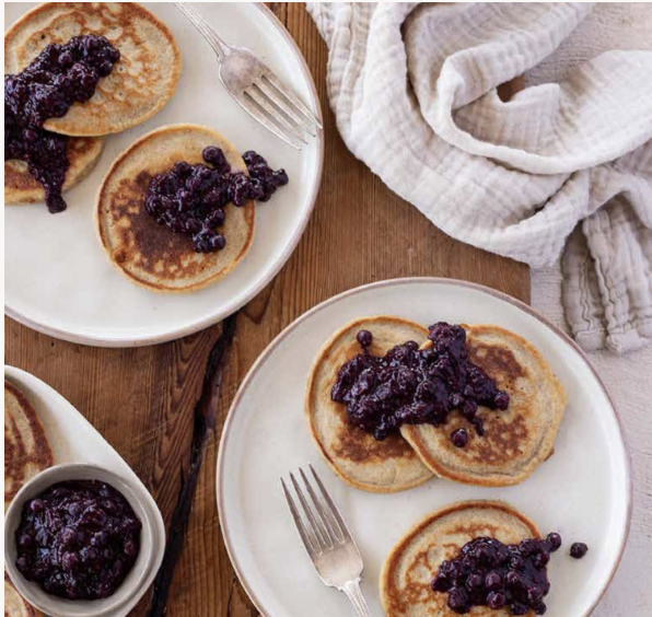 Plate with three small pancakes topped with blueberry jam, fork, and a bowl of blueberry jam on a wooden table.