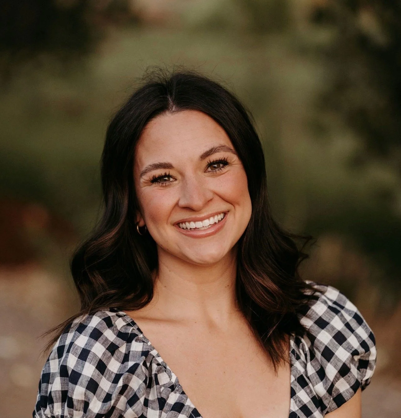 A smiling woman with black hair, wearing a black and white checkered top, standing outdoors with blurred greenery background.