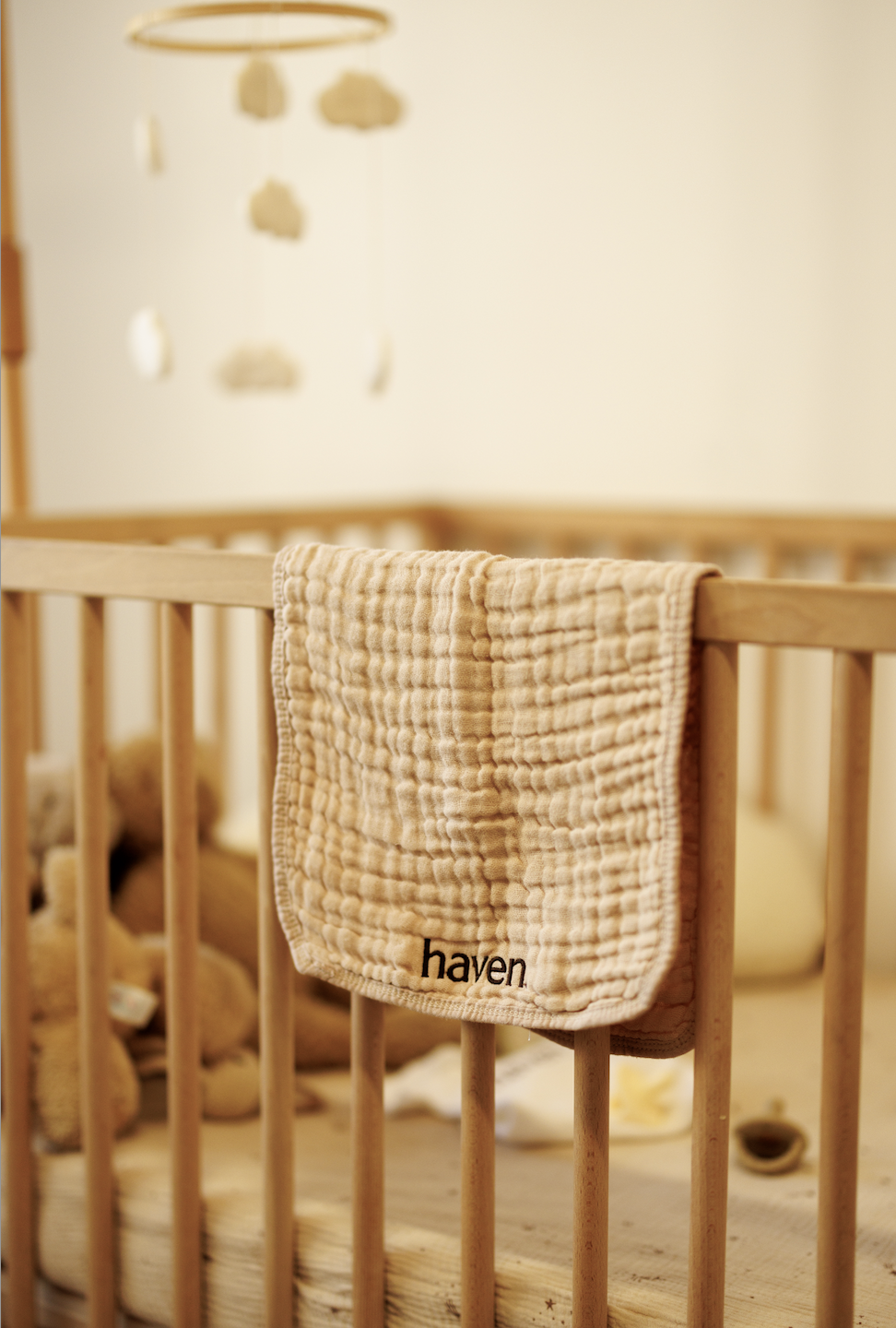 A beige quilted hand towel with the word "haven" embroidered on it, draped over the railing of a wooden crib in a nursery.