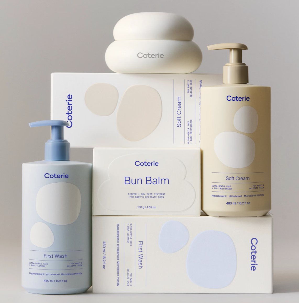 Collection of baby skincare products, including Coterie First Wash, Soft Cream, Bun Balm, and a soap bar, arranged neatly on a gray background.