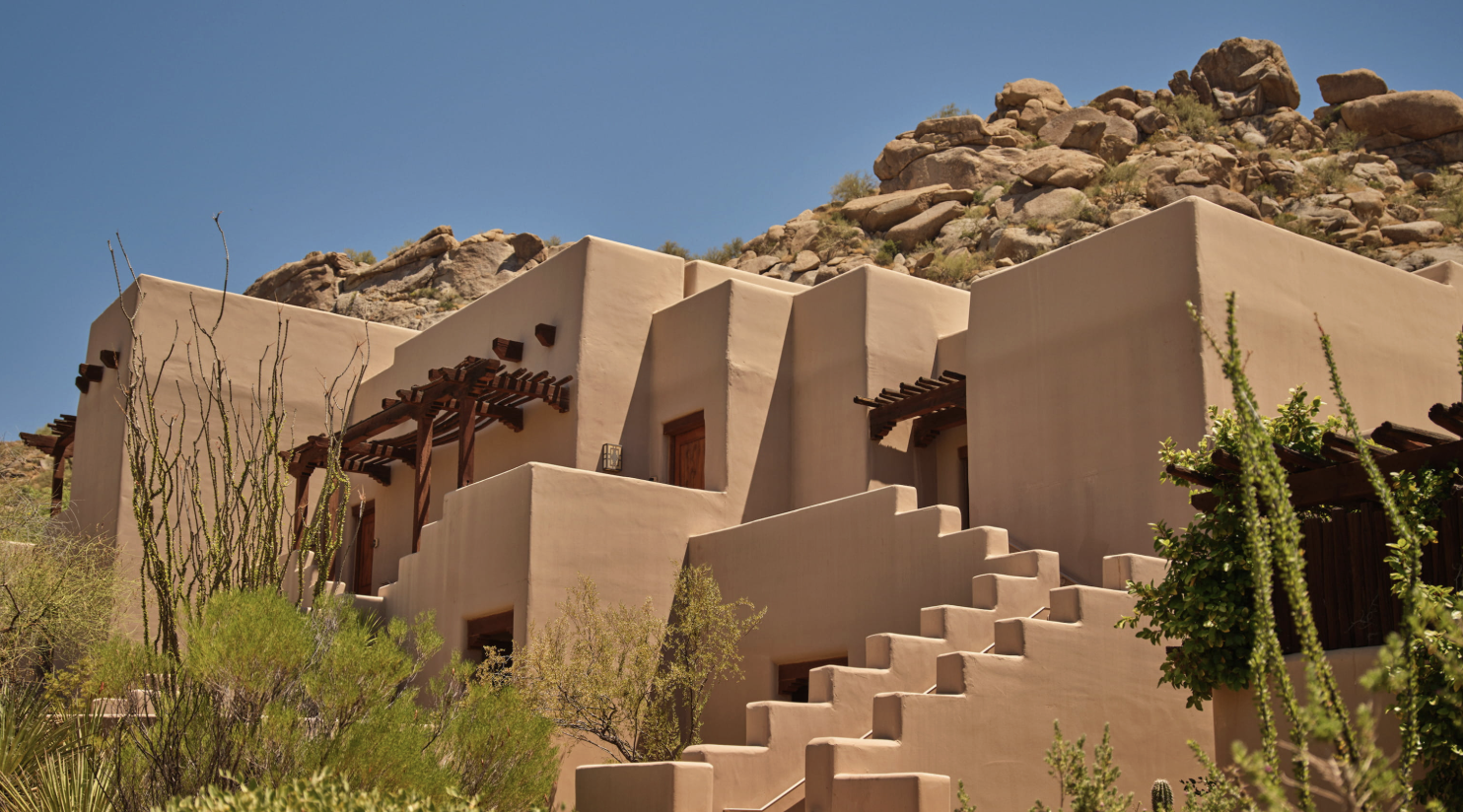 Adobe-style house with multiple levels, wooden pergolas, desert plants, and rocky hillside in background under blue sky.