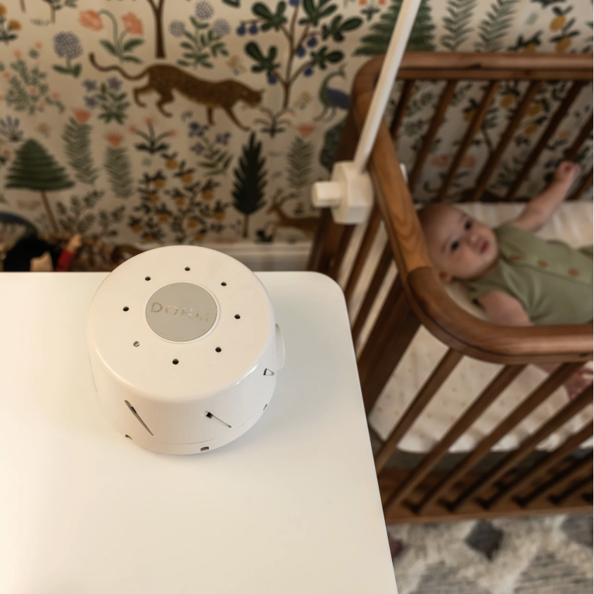 A white, round diffuser on a white table in the foreground with a baby in a crib looking up in the background.