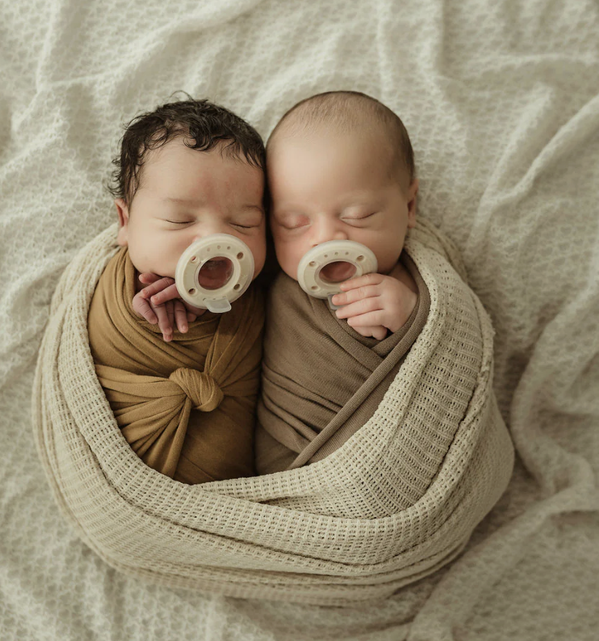 Two swaddled babies sleeping side by side, each with a pacifier in their mouth, wrapped in a cream-colored blanket.