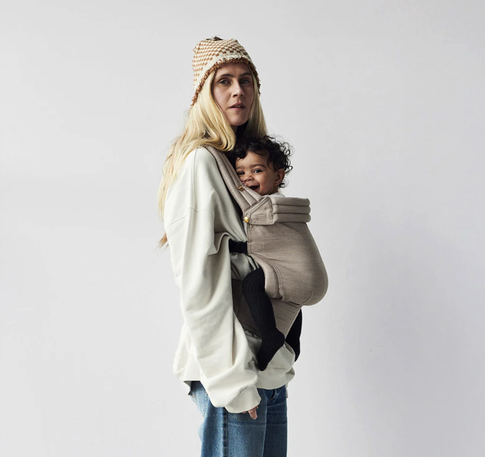 A woman with long blonde hair wearing a beige beanie hat, a cream-colored oversized sweatshirt, and blue jeans, carrying a baby in a tan baby carrier. The baby has curly black hair, a big smile, and is dressed in a black outfit with black socks. The 