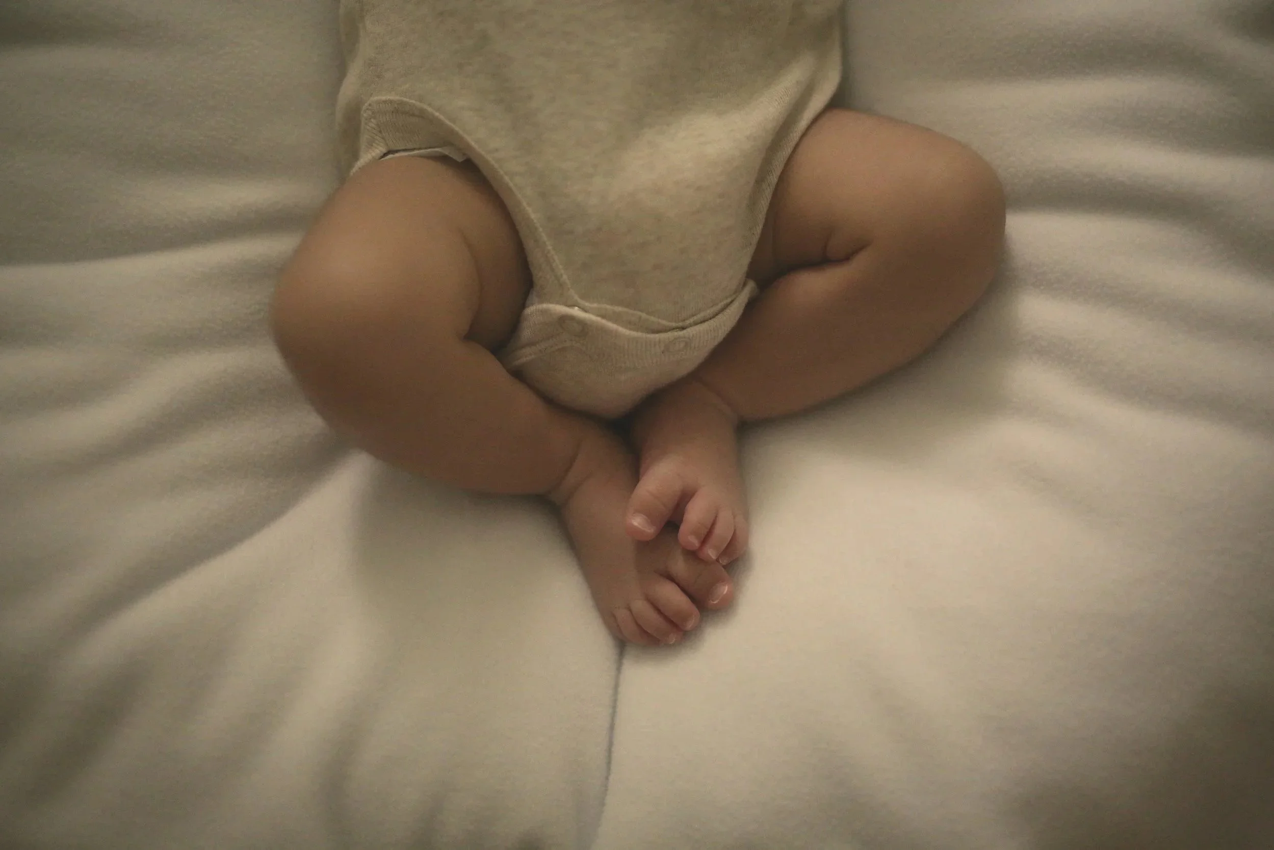 Close-up of a baby sitting on a white bed, wearing a beige onesie.