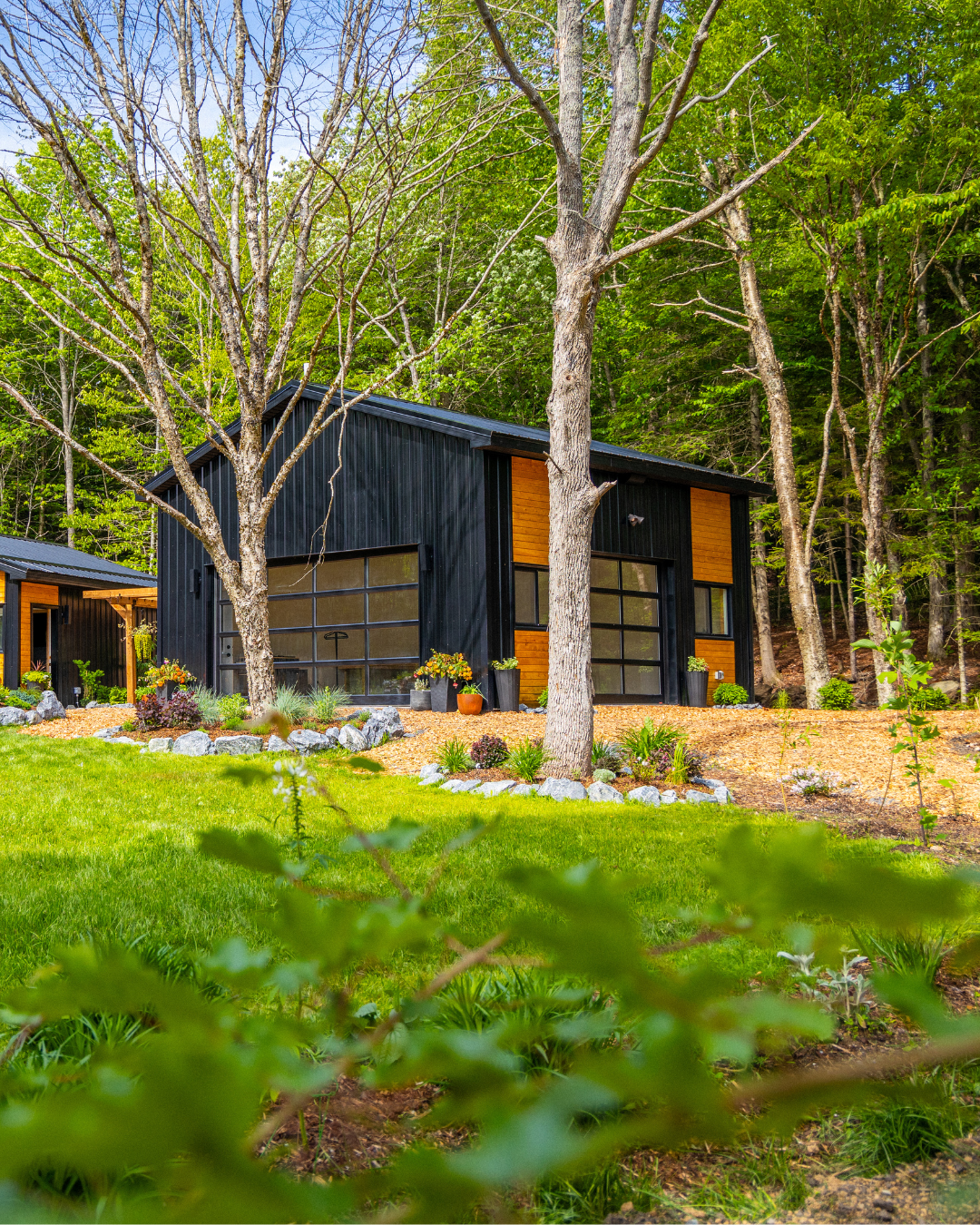 Modern black house with large glass garage doors, surrounded by trees and greenery in a landscaped yard.
