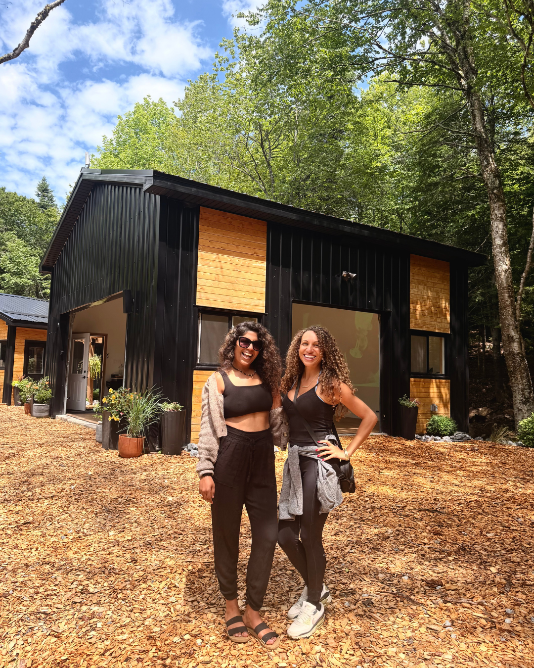 Two women smiling and posing in front of a modern house with black siding and wooden accents, surrounded by trees and potted plants.