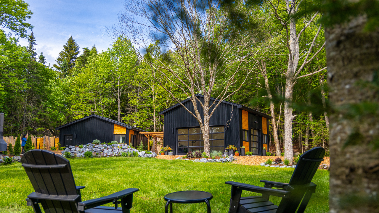 Two black modern houses with large glass windows in a lush green backyard with trees and a flowered landscape, under a partly cloudy sky.