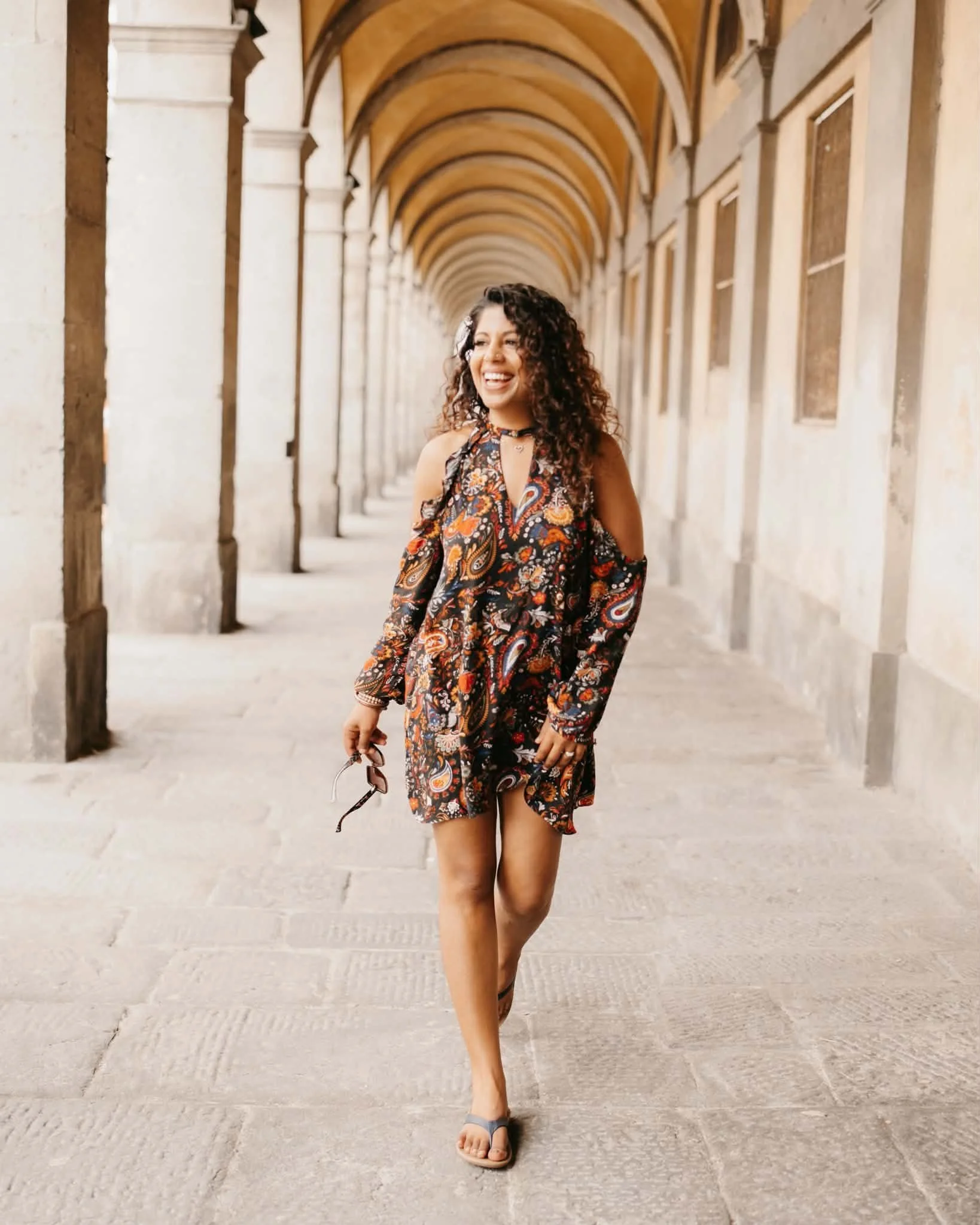 A woman with curly hair, wearing a floral dress, smiling and holding sunglasses, walking through a covered stone corridor with arches.