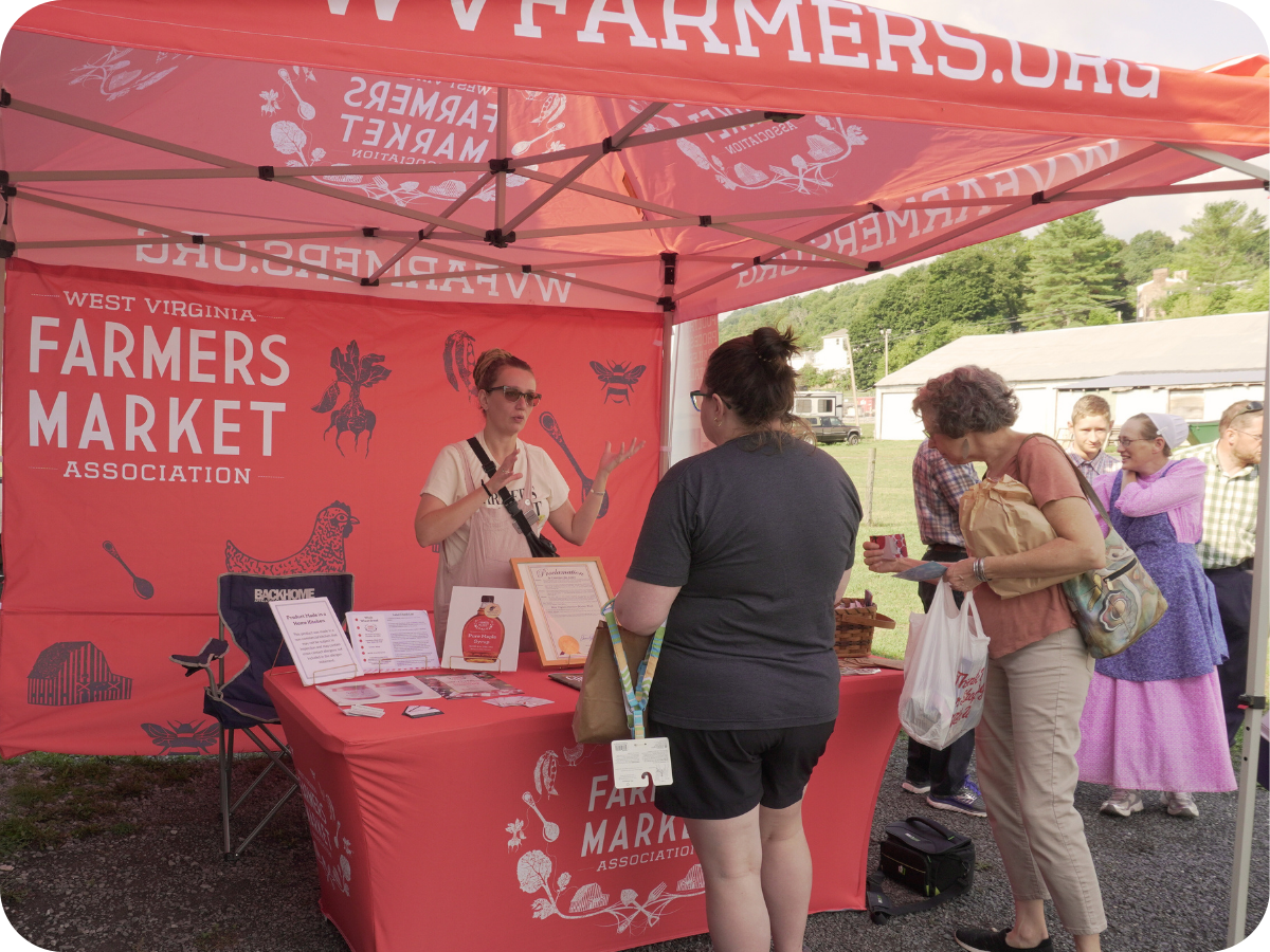A farmer's market booth under a pink canopy with the West Virginia Farmers Market Association logo. Two women are talking to a vendor at the booth, while other people stand in line or browse nearby outdoors, with a farm and trees in the background.