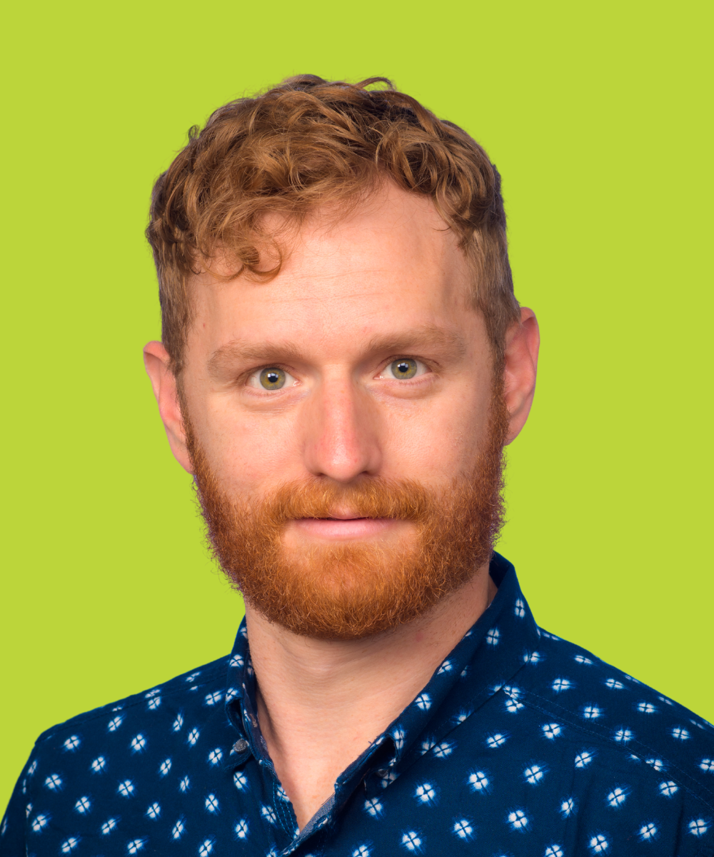 Headshot of a red-haired man with a beard wearing a blue patterned shirt, against a bright green background.