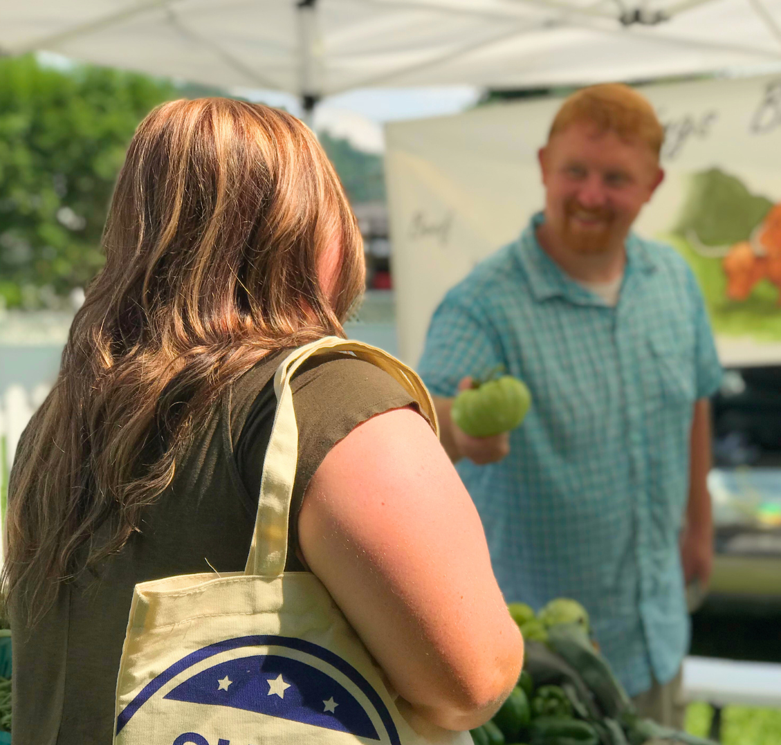 A woman with brown hair carrying a tote bag shopping at a farmers market where a man in a light blue plaid shirt with red hair is holding green bell peppers and smiling.