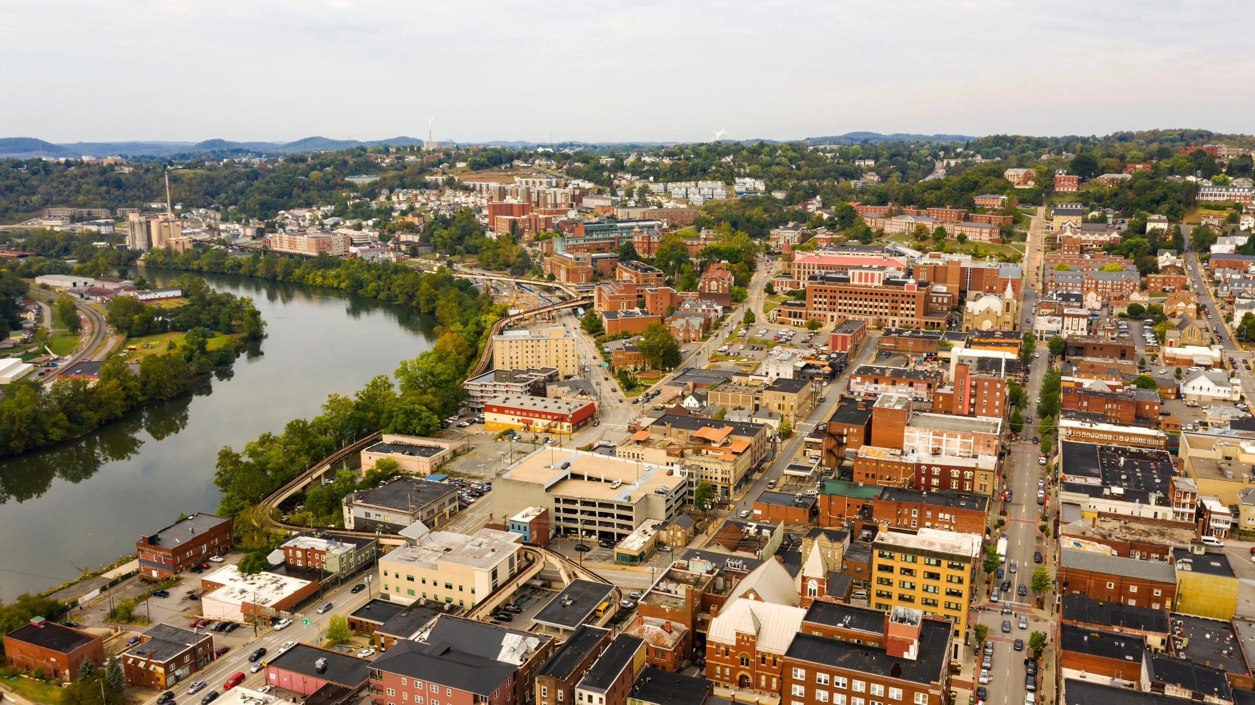 Aerial view of downtown Morgantown, WV. The city is surrounded by buildings, streets, and greenery, with hills in the background.