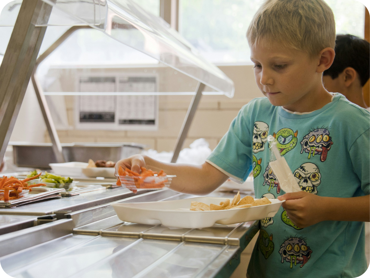 A young boy with blond hair wearing a blue shirt with cartoon characters is serving himself food at a cafeteria-style food line. He is holding a tray using tongs to pick up carrots or similar vegetables from a serving station.