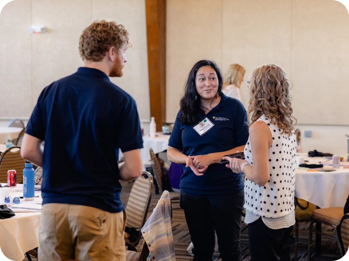 Three people are talking in a conference room with tables and chairs. The woman in the middle is wearing a dark shirt and the women on the right is wearing a white polka dot blouse.