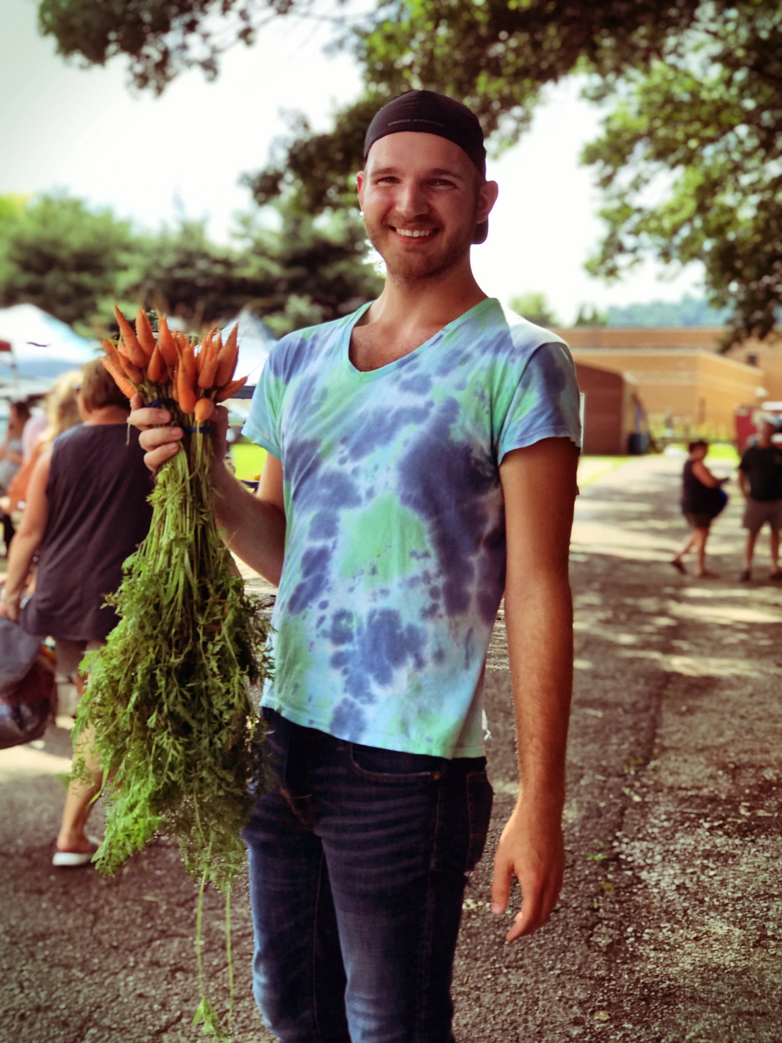 Smiling man in a tie-dye shirt holding a bunch of carrots at an outdoor market.