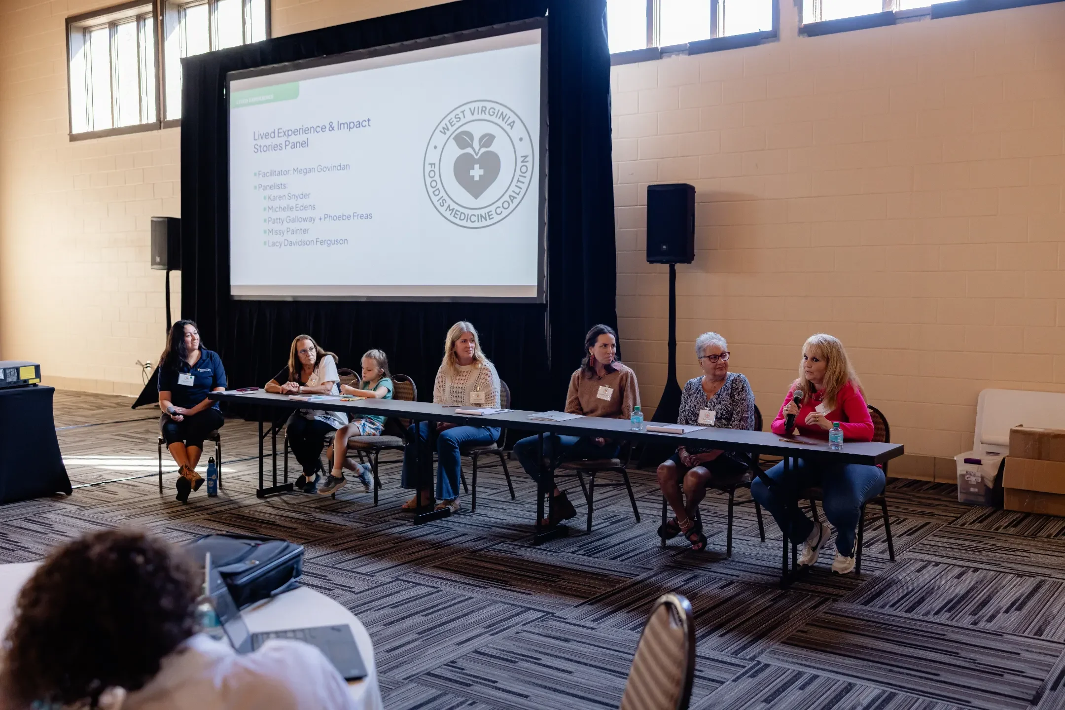 Panel of six women and one young girl sitting at a long table during a conference, with a large screen behind them displaying a presentation titled 'Lived Experience & Impact Stories Panel' and the West Virginia Food & Medicine Coalition logo.