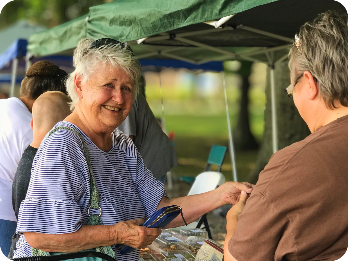 Smiling elderly woman talking to a vendor at an outdoor market stall with tents.