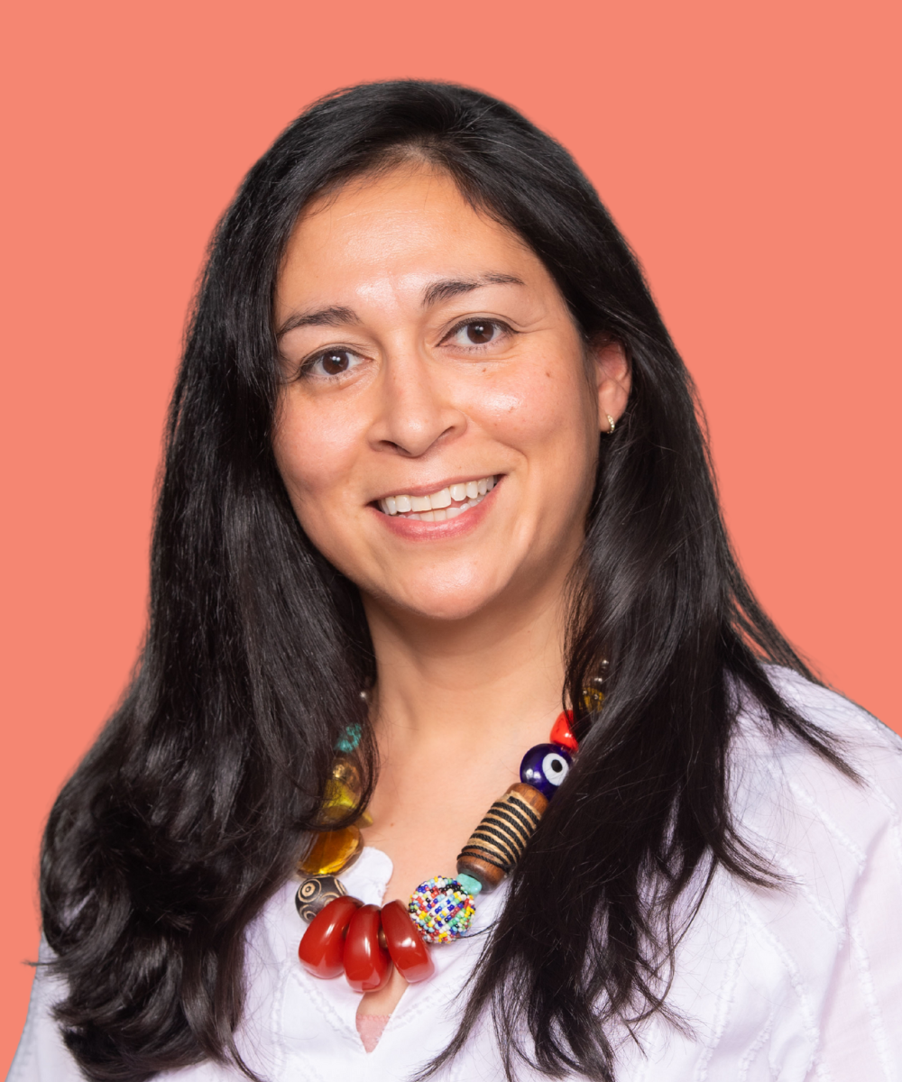 A woman with long black hair smiling against a coral background, wearing a colorful beaded necklace with various patterns and shapes.