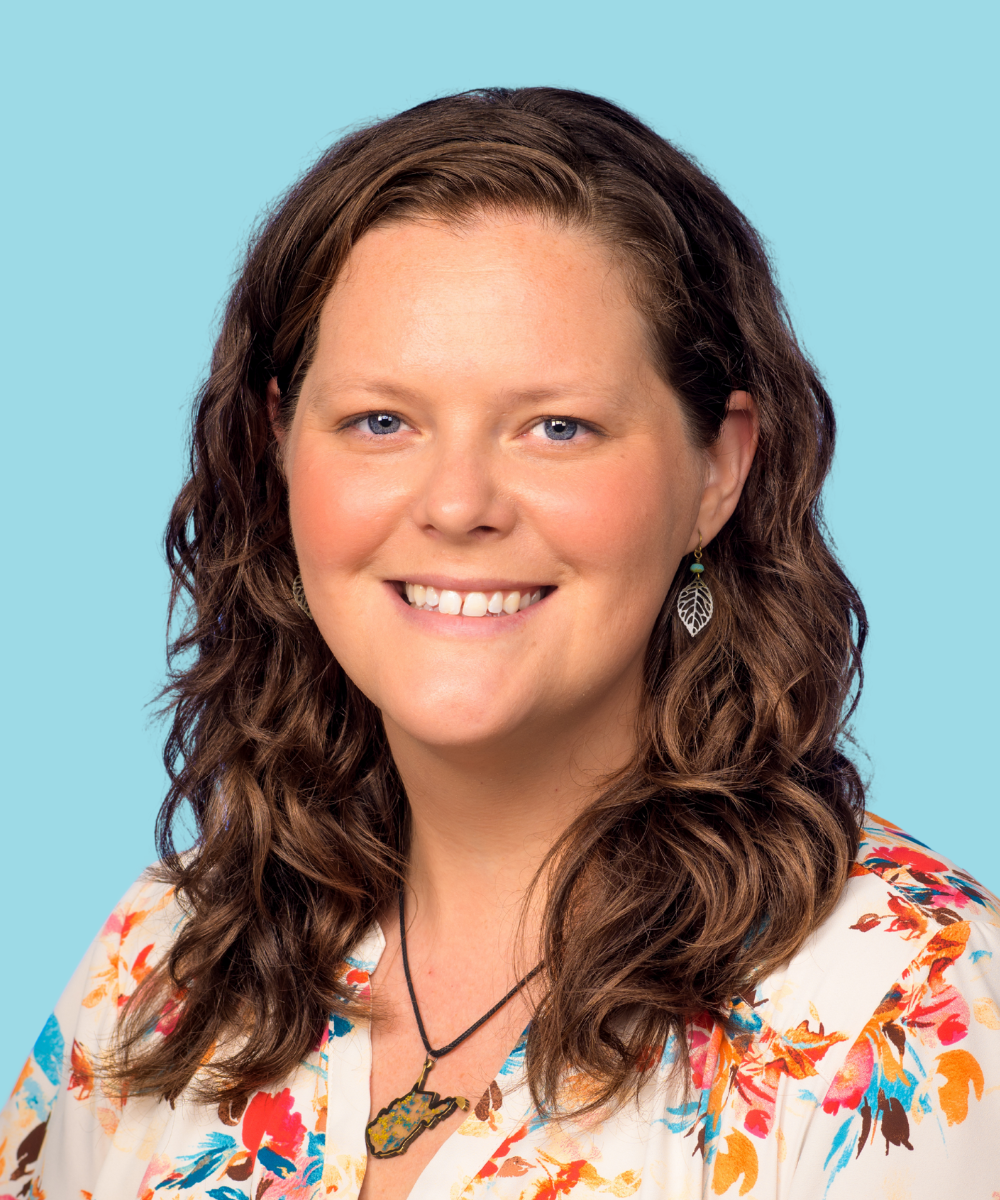 A woman with wavy brown hair, smiling, wearing a floral shirt, leaf-shaped earring, and a necklace pendant in the shape of West Virginia, against a light blue background.