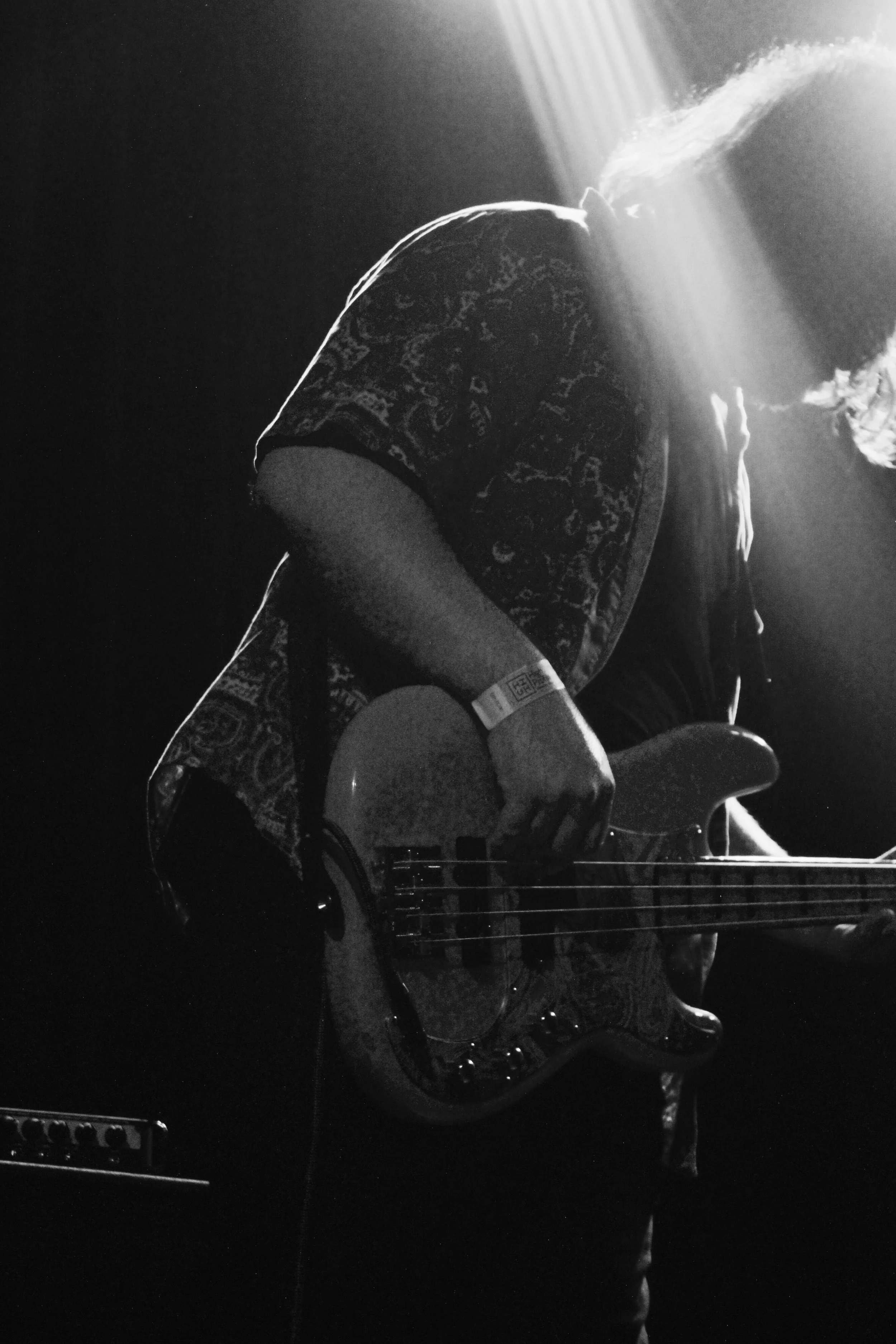 Musician playing an electric bass guitar on stage, backlit with intense light, in black and white.