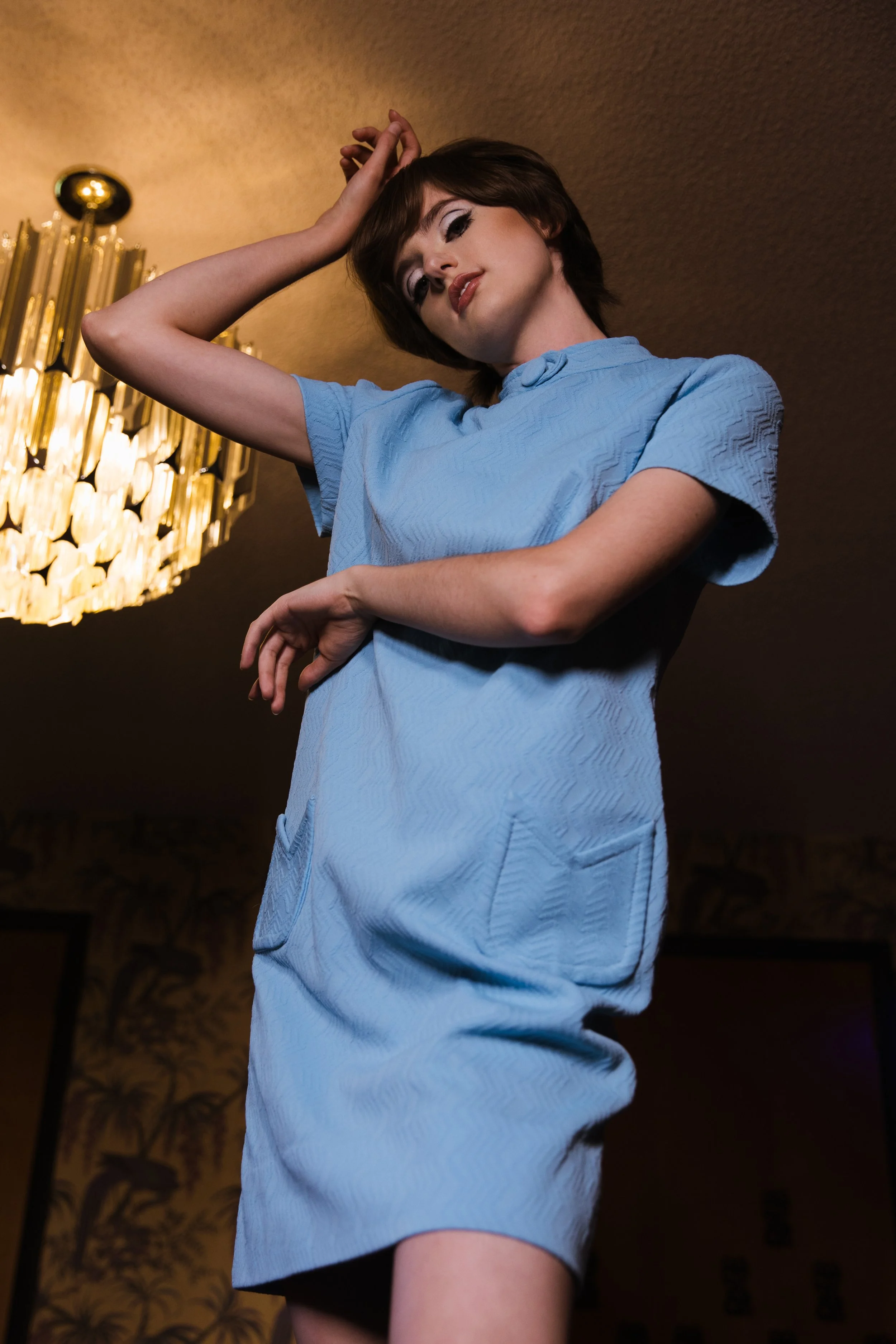 Young woman in a light blue dress posing indoors with a chandelier in the background.