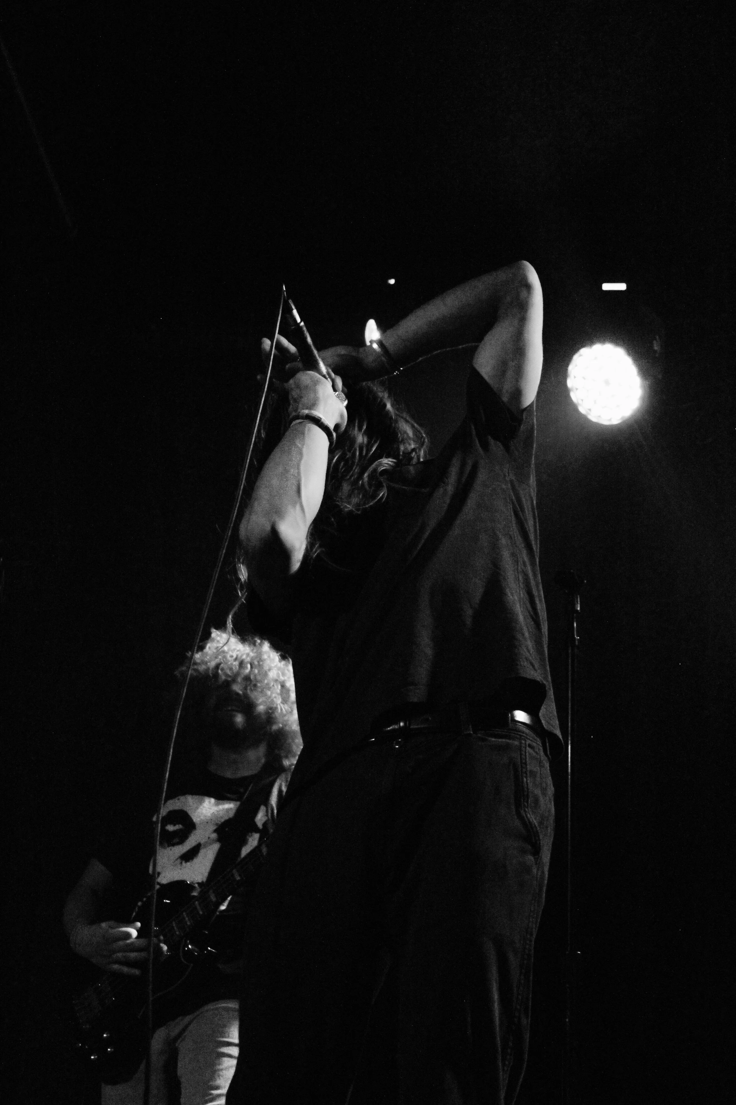A black-and-white photo of a singer performing on stage, holding a microphone with one arm raised, and a guitarist playing in the background.