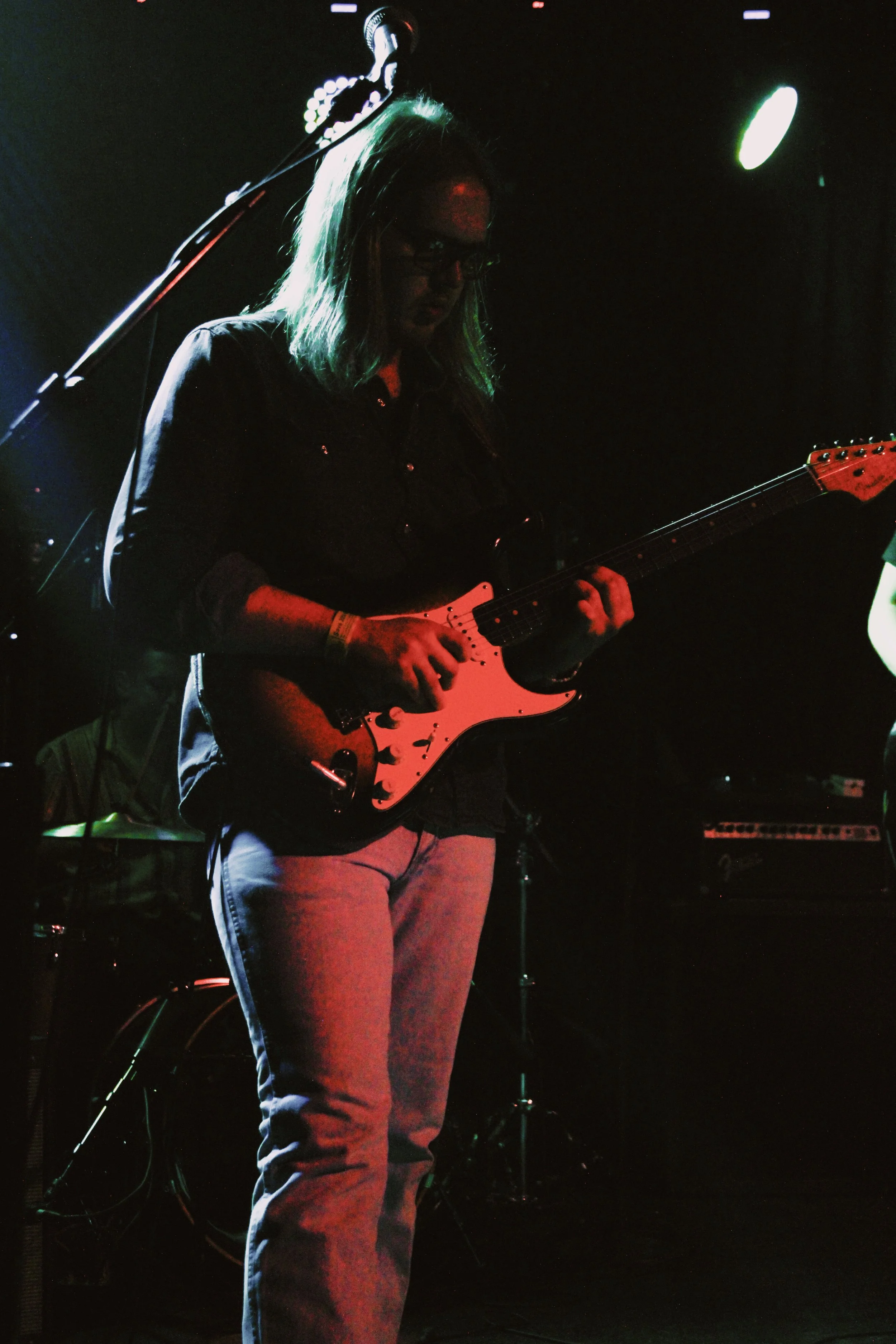 A person playing an electric guitar on stage, with dark lighting and colorful stage lights.
